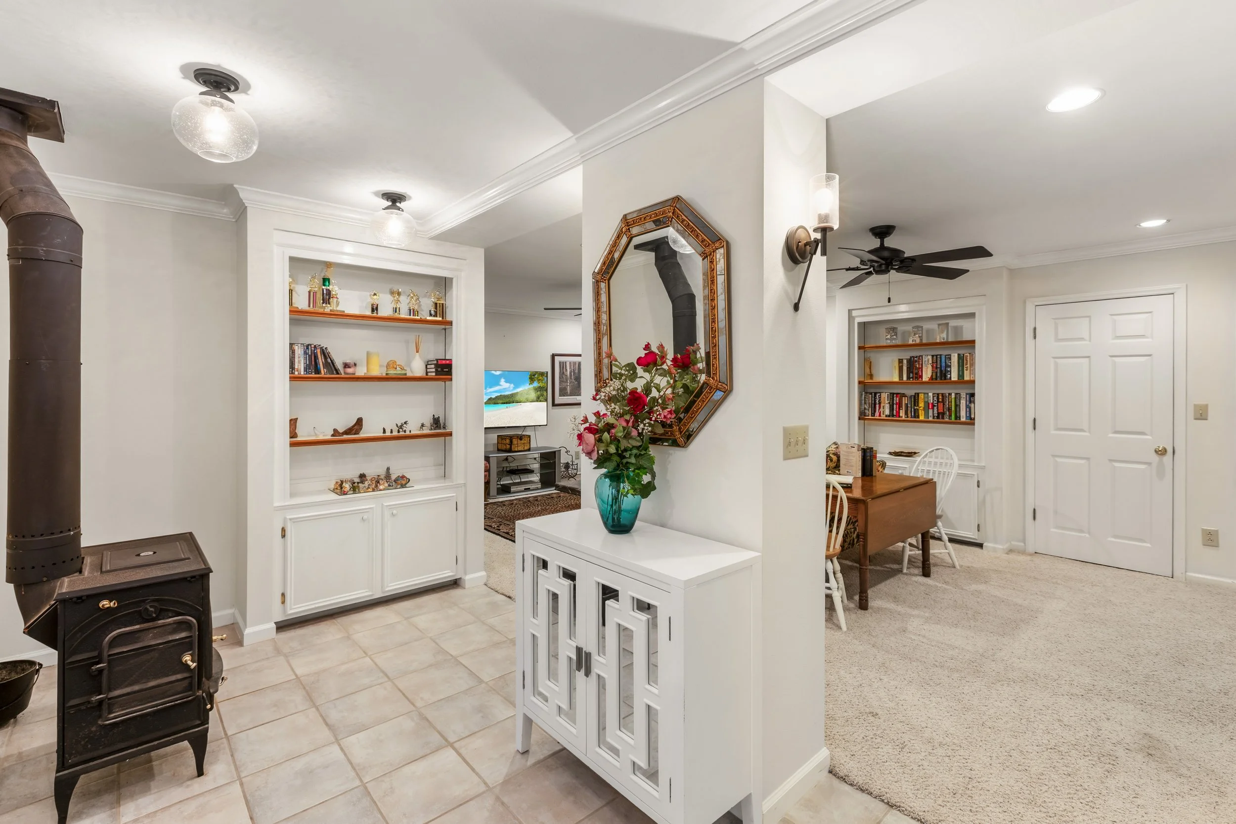 Interior view of a home showing a living and dining area with white walls, built-inbookshelves filled with books and decorative items, a small white cabinet with a flower vase, and a corner of a wood stove with a flue pipe.