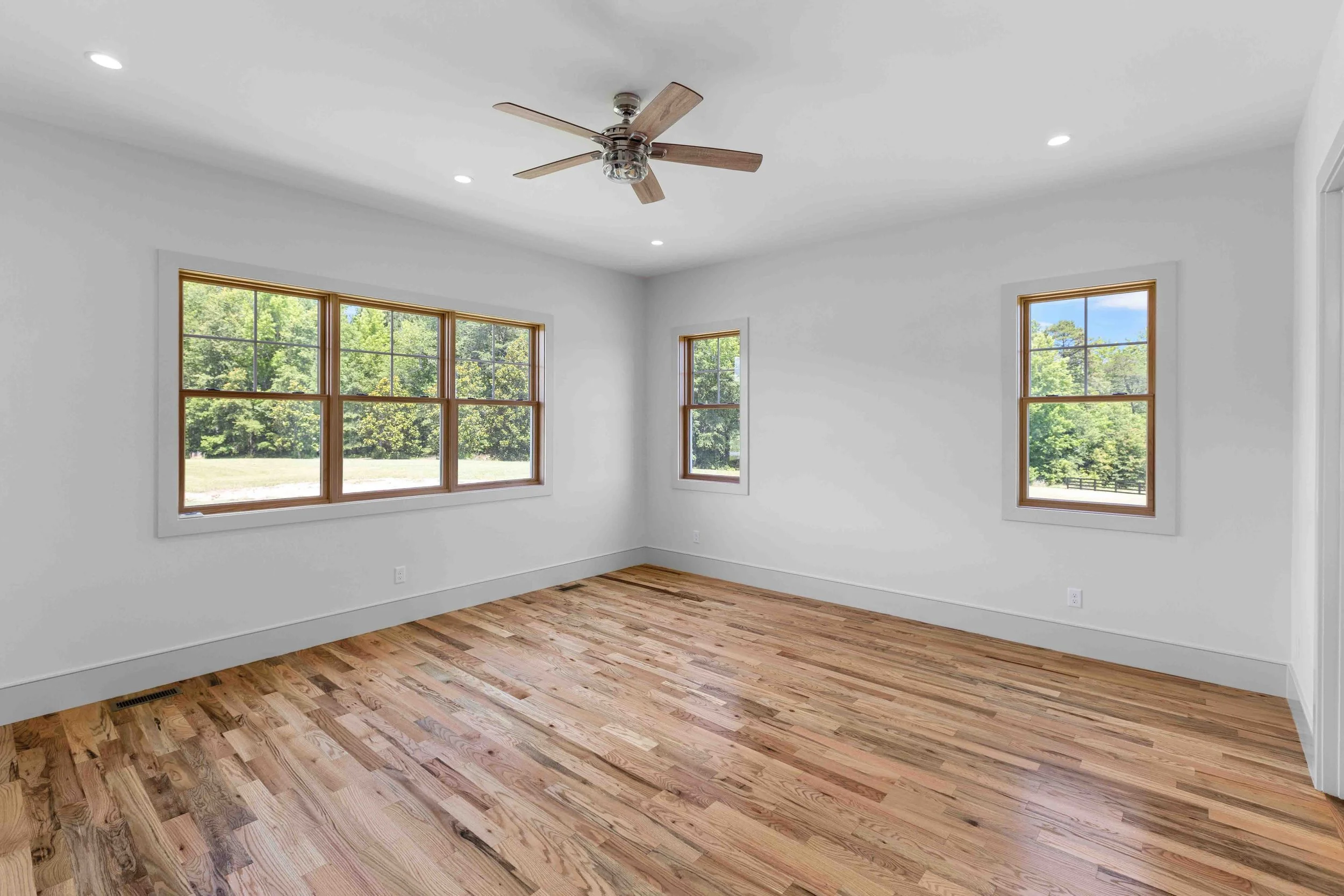 Empty room with white walls, hardwood floors, three windows showing green trees outside, a ceiling fan, and recessed lighting.