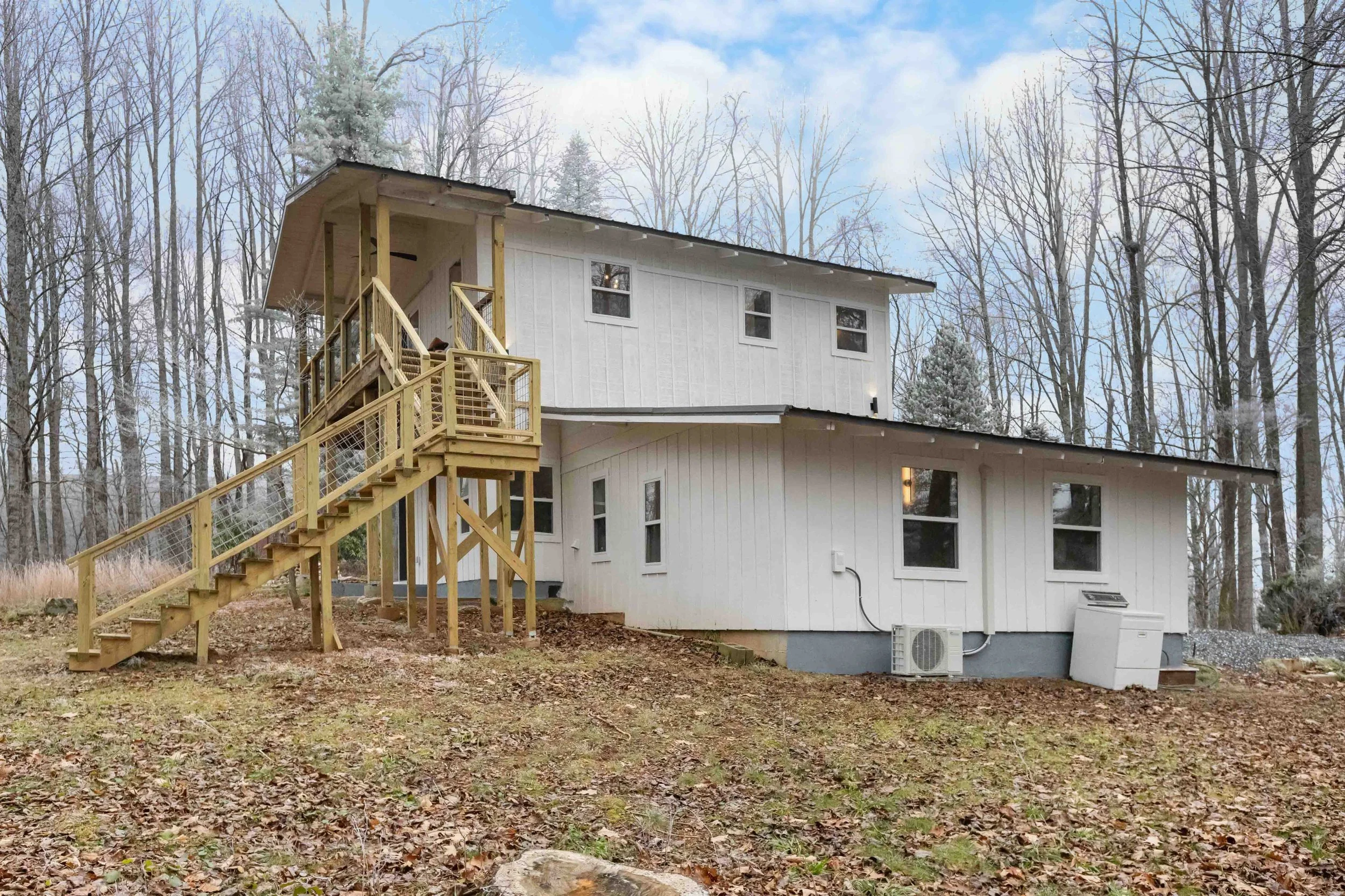 A two-story white house with an exterior staircase on a sloped backyard, surrounded by leafless trees, brown fallen leaves on the ground, and a partly cloudy sky.