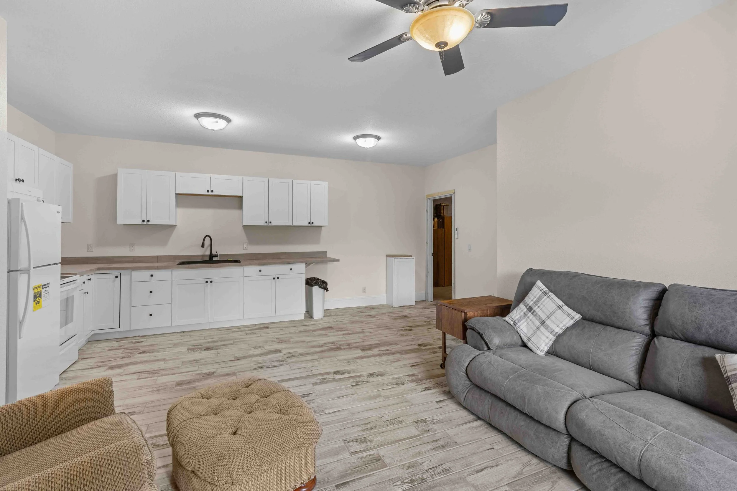 Living room with grey couch and throw pillows, beige armchair, wooden side table, kitchen with white cabinets, countertop, sink, and appliances, ceiling fan, and ceiling lights.