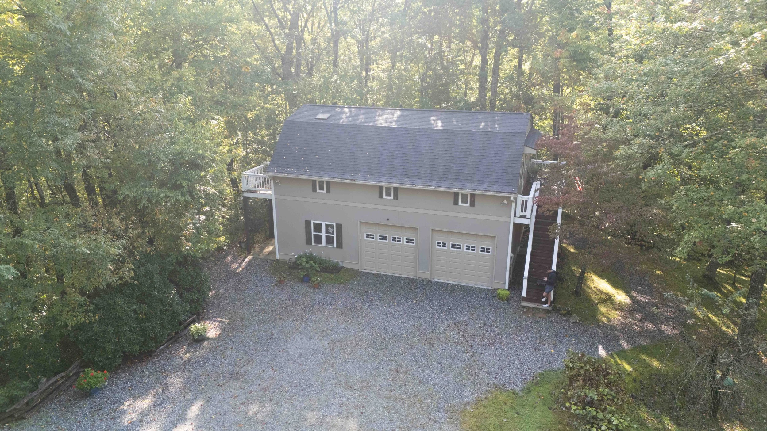 Aerial view of a two-story house surrounded by trees, with a gravel driveway, three garage doors, a deck on the side, and stairs leading up to the deck, with a person standing at the foot of the stairs.