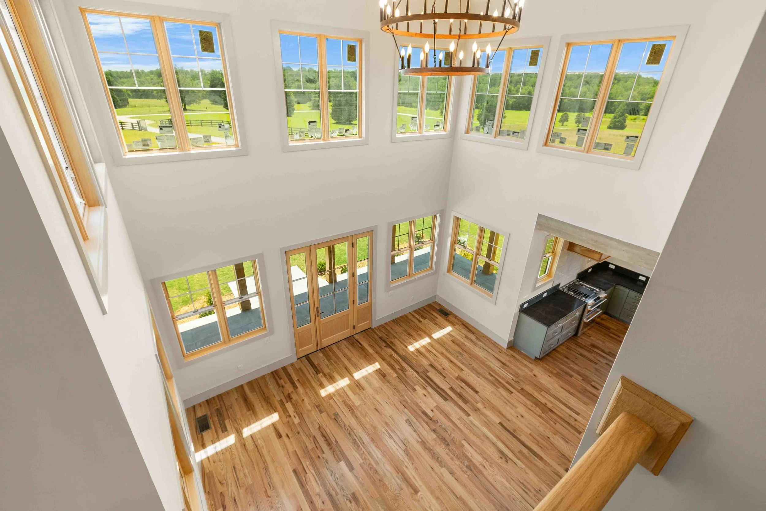 Empty living room with high ceiling, hardwood floors, multiple large windows showing green fields, a wood and metal chandelier, and a kitchen area with gray cabinets and black countertops.