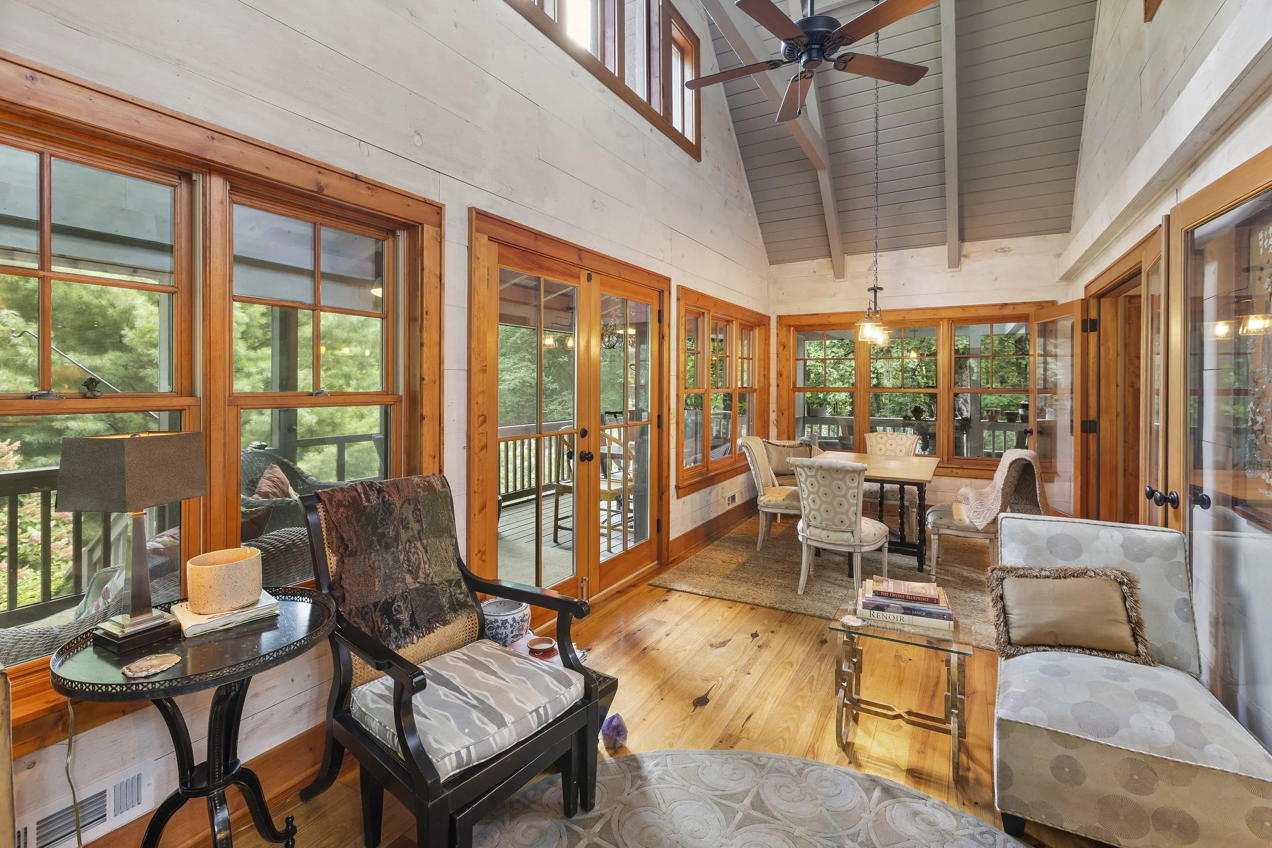 Sunroom with large wooden-framed windows, hardwood floors, and a mix of chairs around a small dining table.