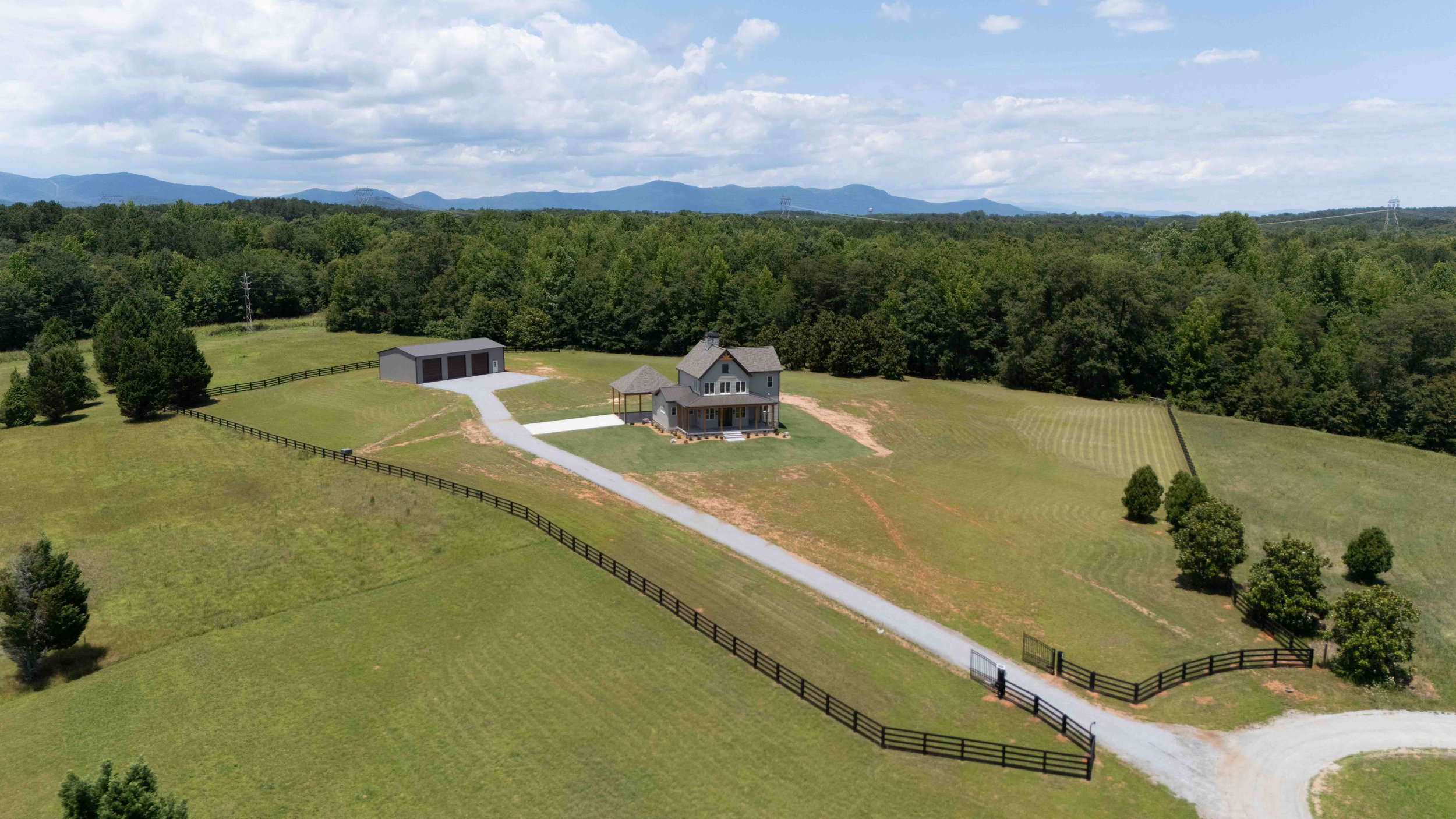 Aerial view of a countryside house surrounded by green lawns and trees, with a gravel driveway leading to a garage and fencing around the property, mountain range in the background under a partly cloudy sky.