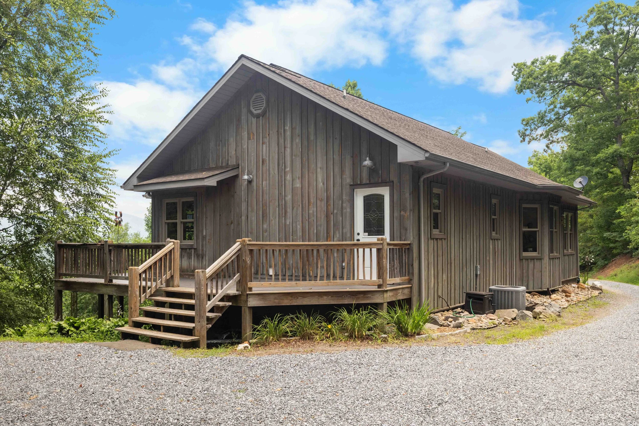 A rustic wooden house with a deck and stairs, set on a gravel driveway surrounded by green trees and a blue sky.