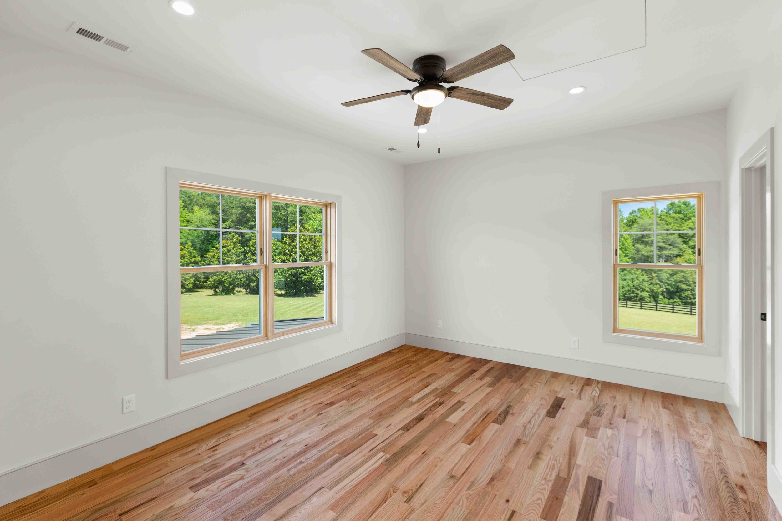 Empty room with white walls, hardwood floor, two windows showing green trees outside, ceiling fan, and ceiling lights.