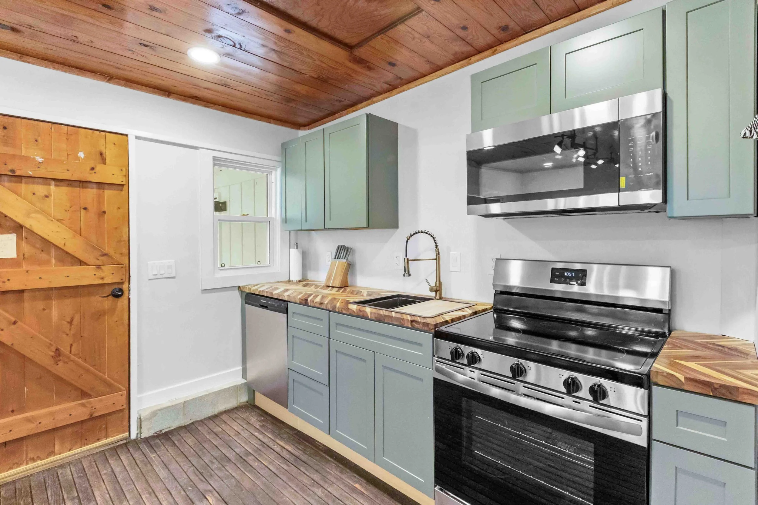 Kitchen with light green cabinets, wooden countertops, stainless steel stove and microwave, white walls, wooden ceiling, and a small window.