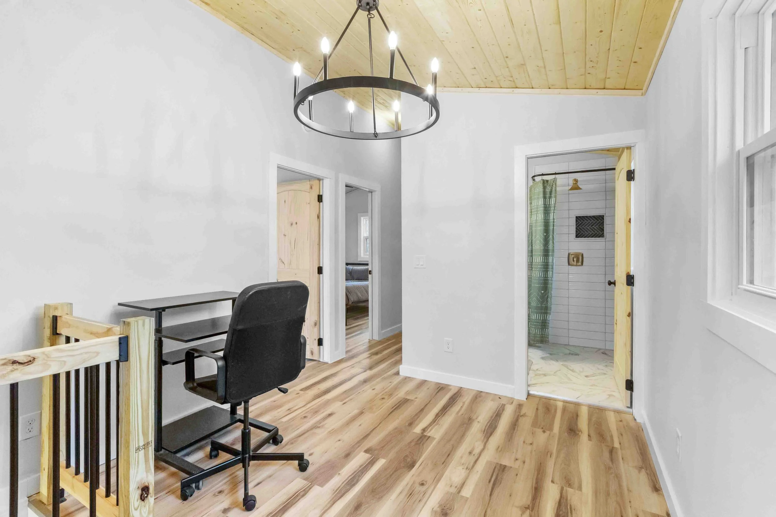 Interior of a room with a black desk and chair near a staircase, wood floors, white walls, a black chandelier ceiling light, and doorways leading to other rooms and a bathroom with a shower.