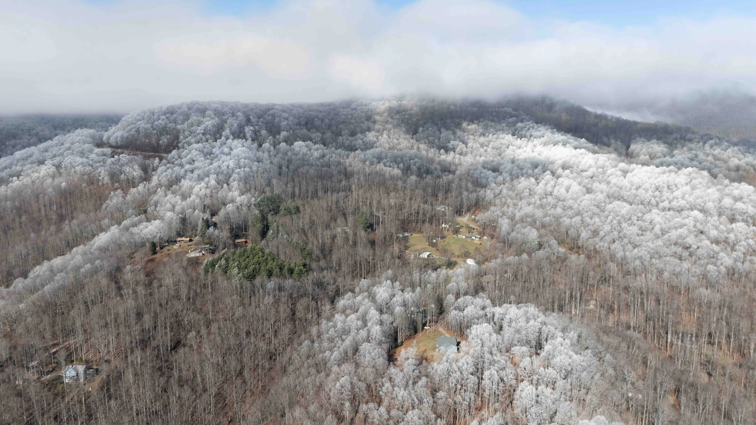 Aerial view of a winter landscape with snow-covered trees on rolling hills, with some houses and roads visible among the trees, and low clouds in the background.