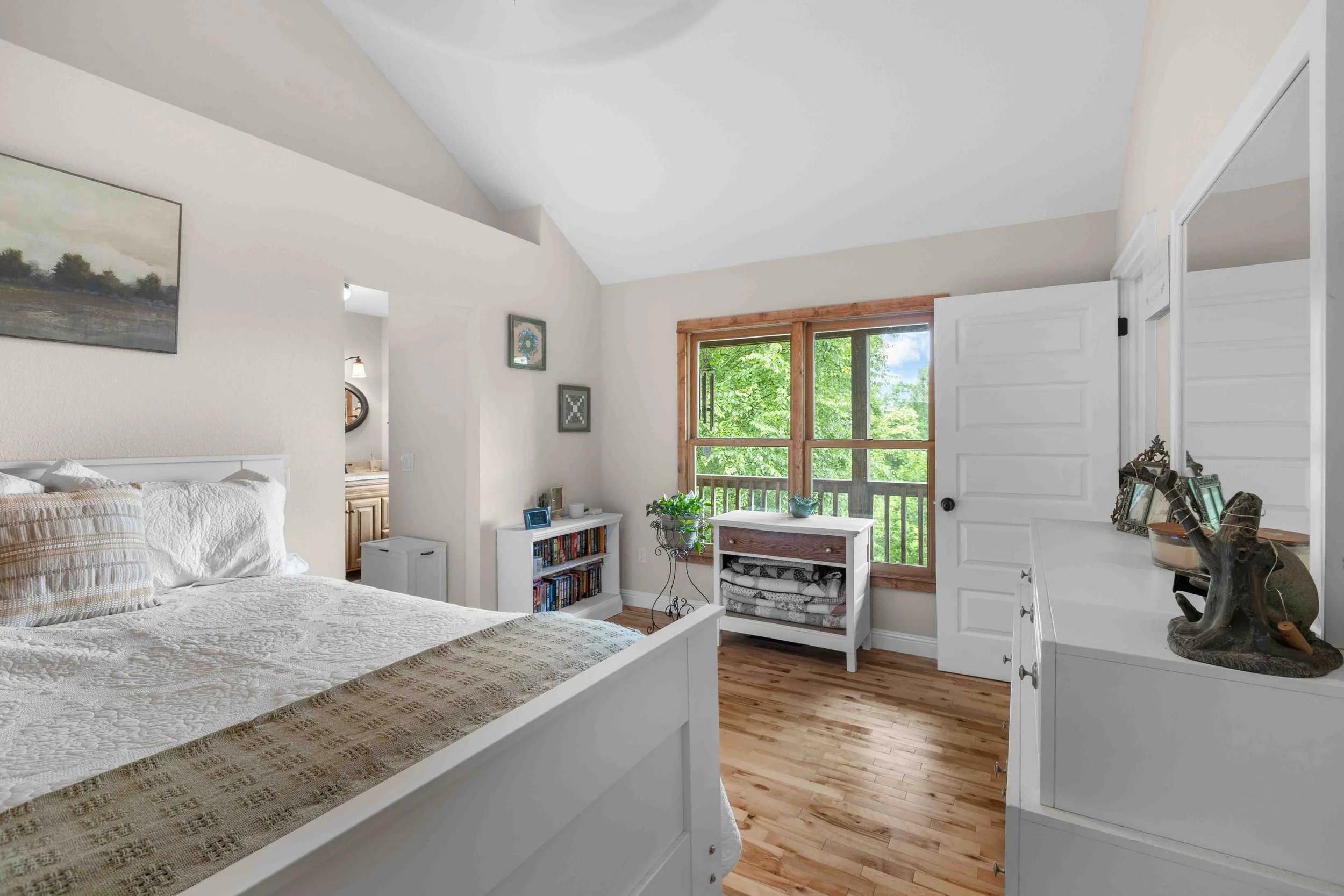 Bright bedroom with white furniture, wooden floors, and a large window showing green trees outside.