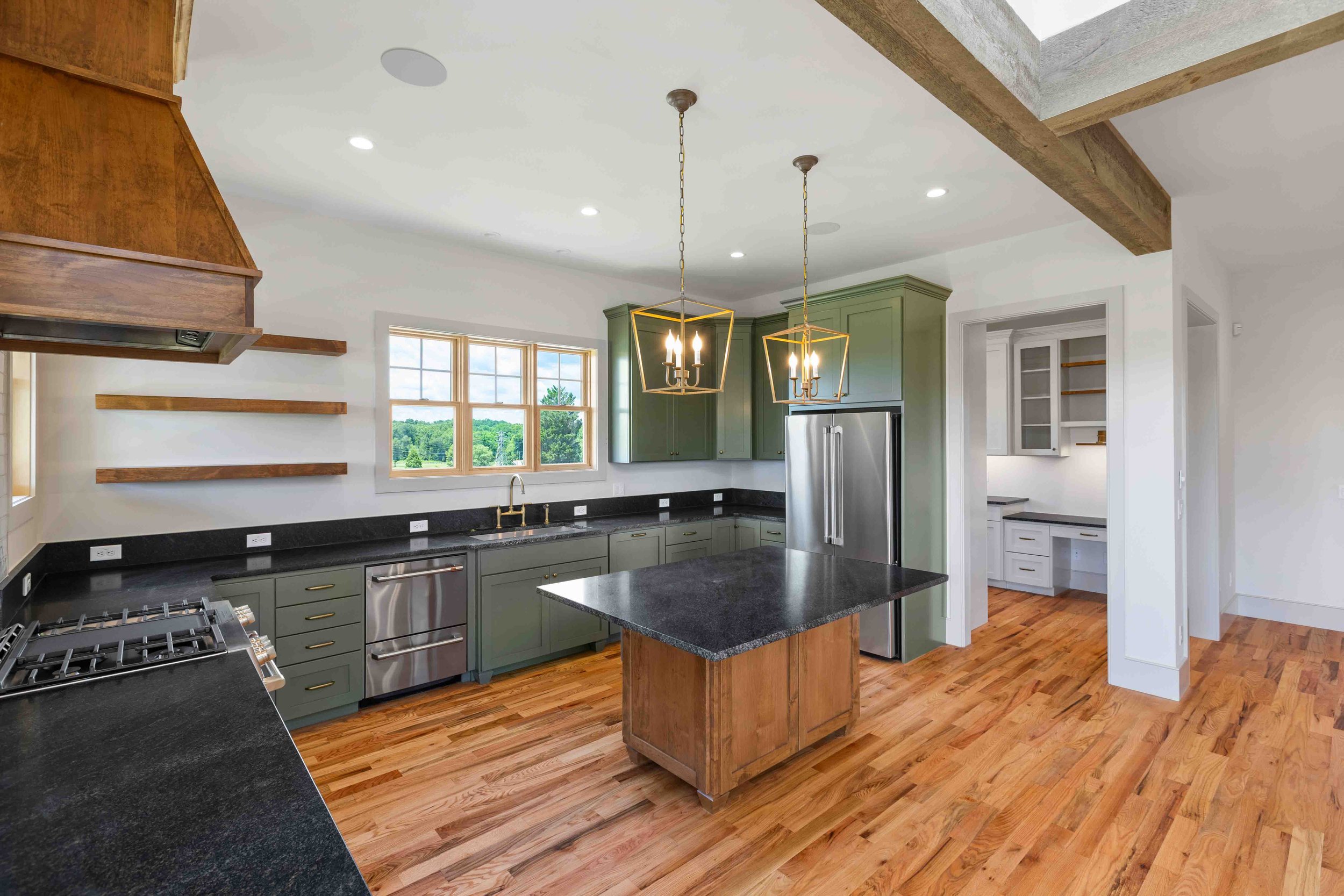 Modern kitchen with green cabinets, black countertops, hardwood floors, and wooden open shelving. Features two pendant lights above a central island and stainless steel appliances, with a view of trees through the window.