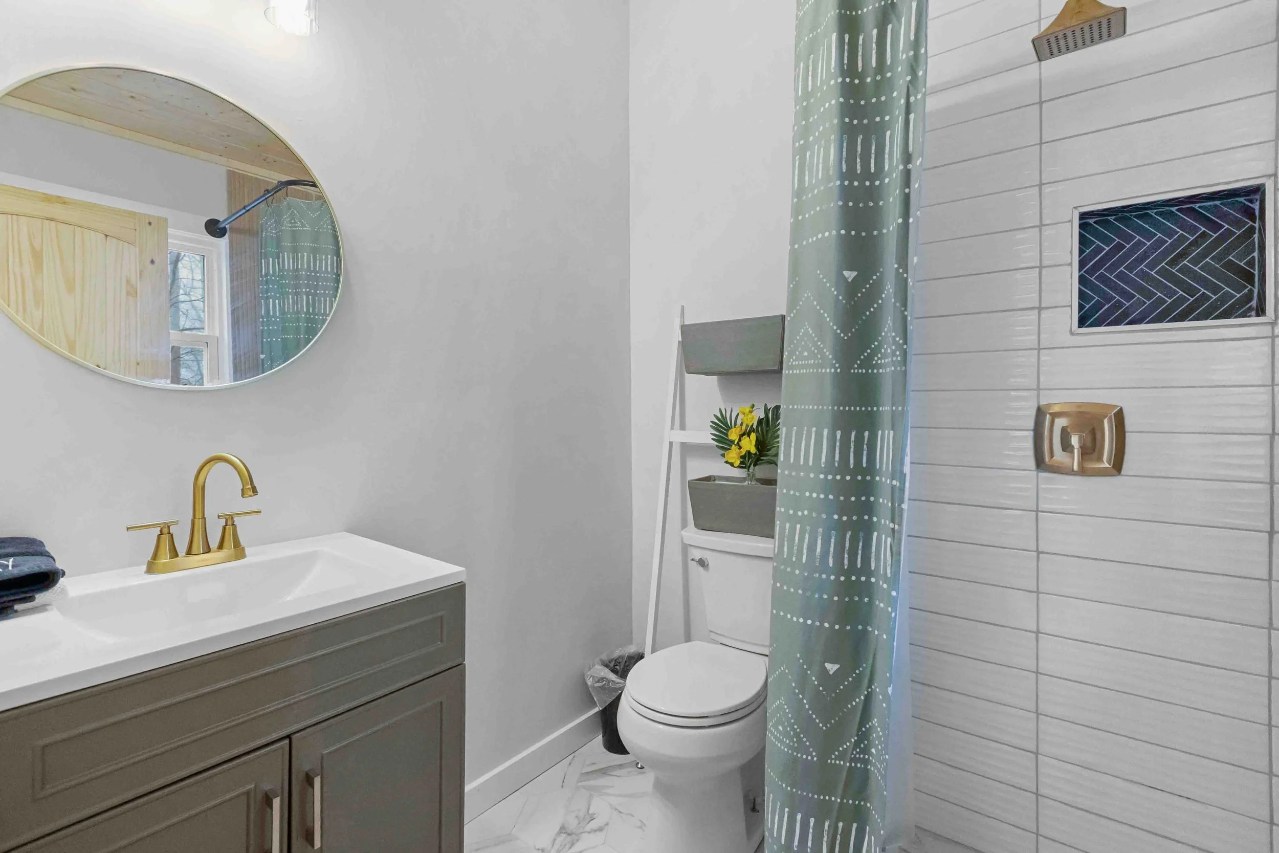Modern bathroom with a white vanity, gold faucet, and an oval mirror. There is a toilet with a gray shelf holding yellow flowers. A shower with a patterned curtain, tiled walls, and a built-in niche for toiletries.