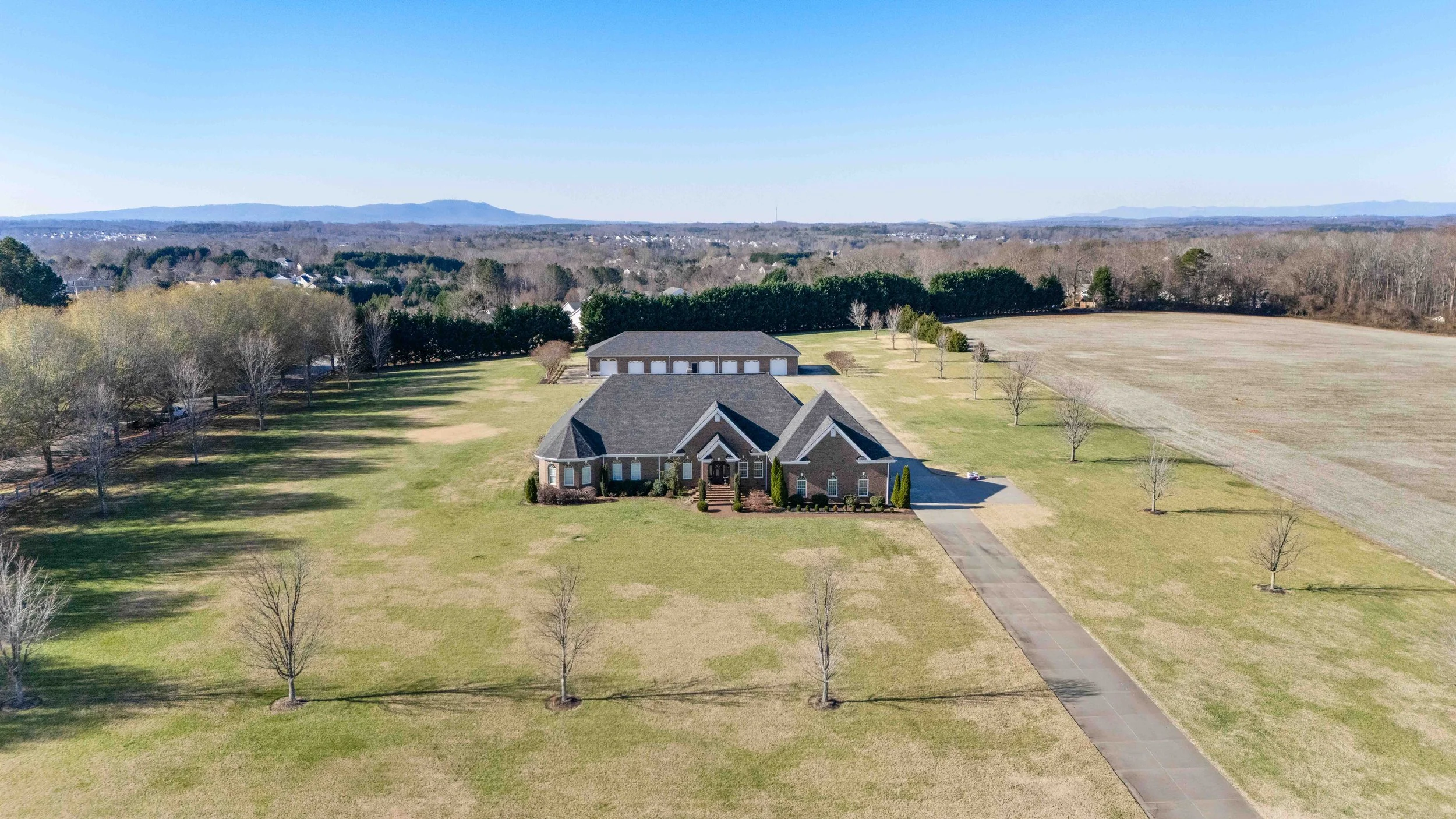 A large brick house with a gray roof on a spacious lawn, lined with leafless trees, with a driveway leading up to it. There is a detached garage or barn in the background, and open fields extend around the property with distant hills on the horizon.