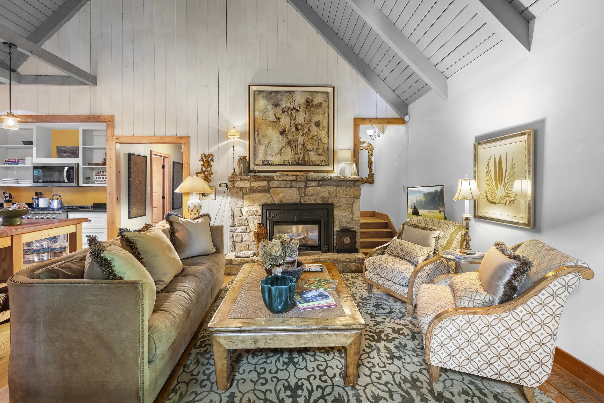 Living room with a stone fireplace, beige and patterned armchairs, a wooden coffee table with magazines, decorative flowers, and various lamps. Open kitchen area visible in the background.