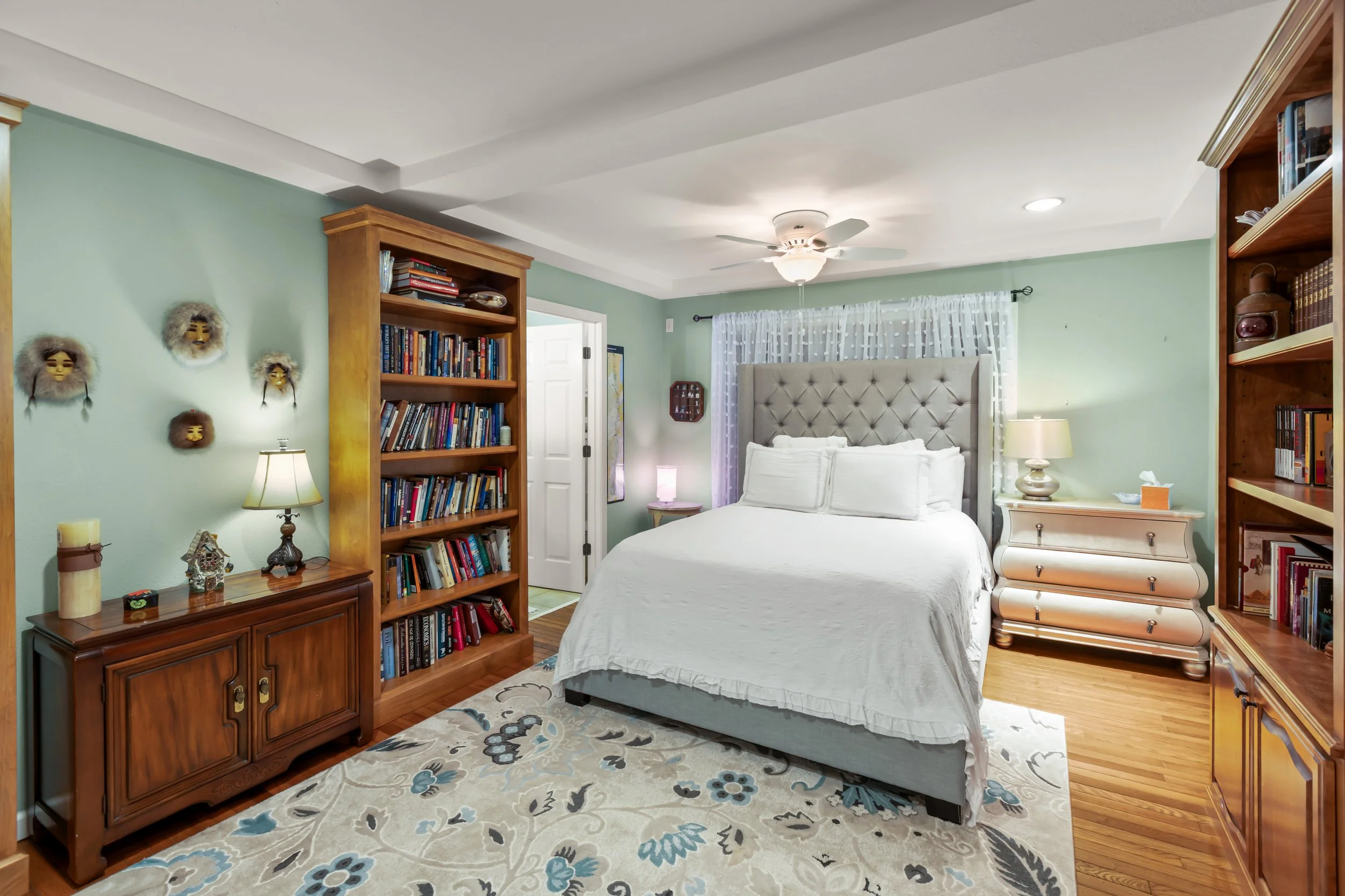 Bedroom with a large bed, twin nightstands with lamps, wooden bookshelves filled with books, wooden cabinet, decorative wall masks, ceiling fan, hardwood floors, floral area rug, light green walls, and white curtains.