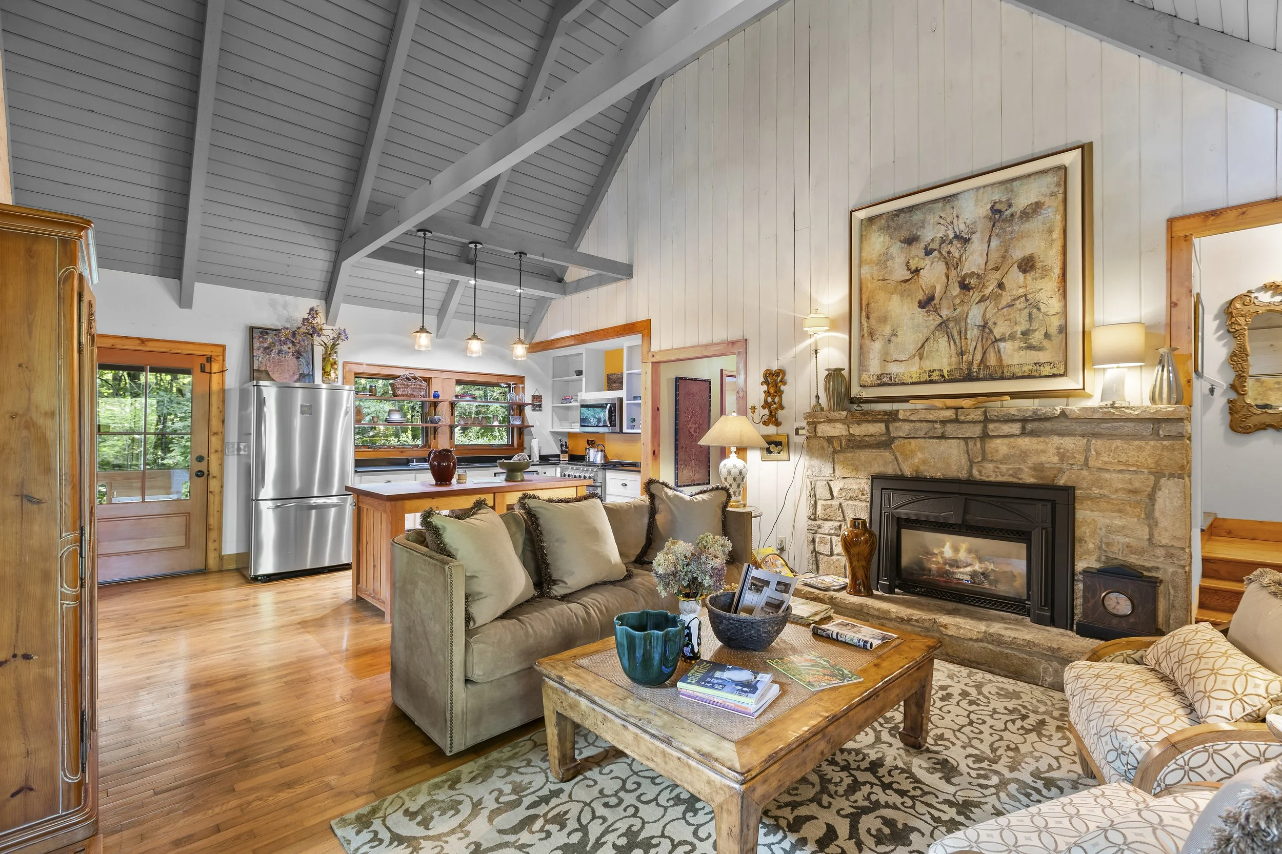 Living room with a stone fireplace, beige sofa, patterned armchair, wooden coffee table, and a yellow wood-paneled wall with large artwork. Open kitchen in background with stainless steel refrigerator, wooden island, and windows.