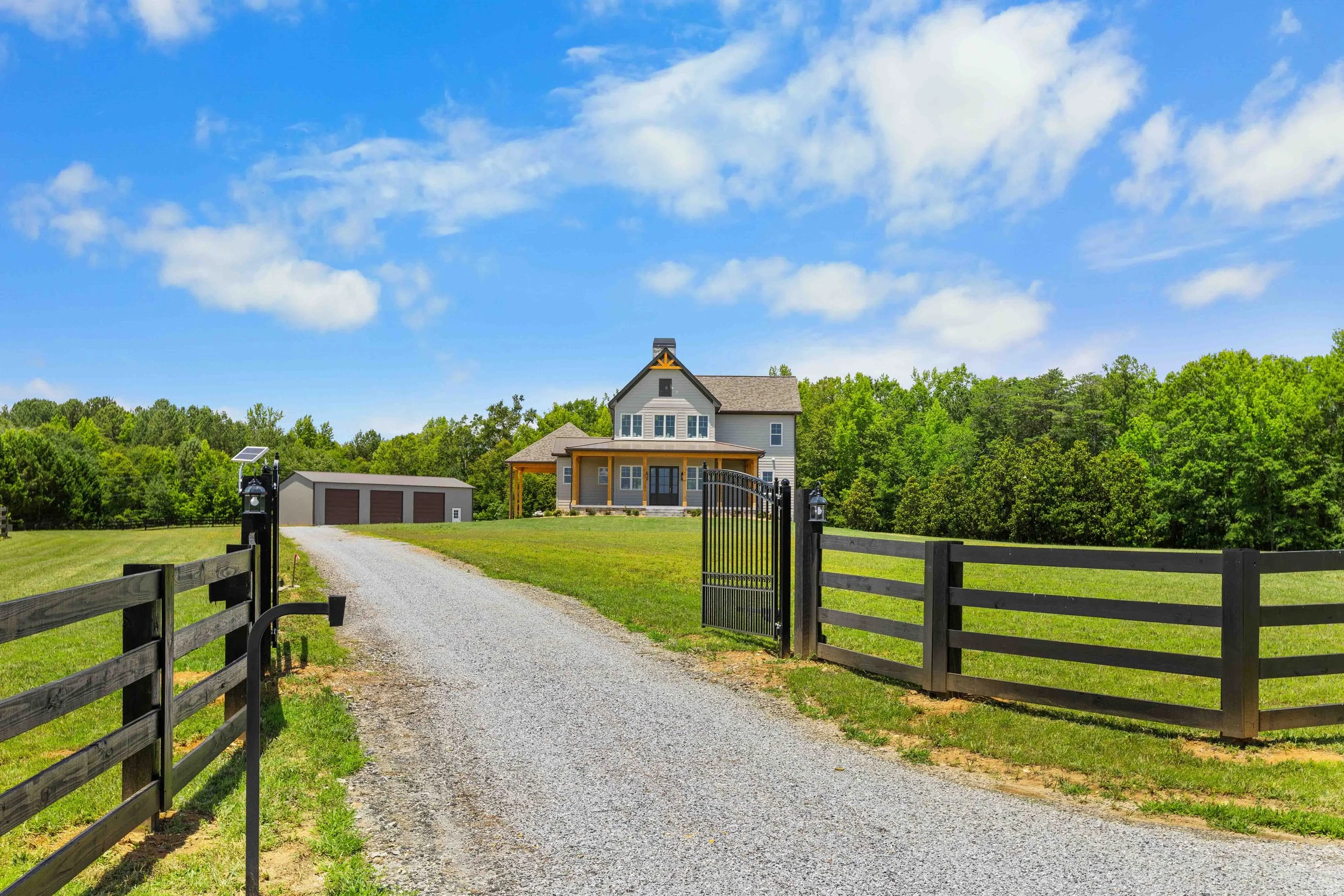 Gravel driveway leading to a house with a covered porch and a detached garage, surrounded by green grass and trees under a partly cloudy blue sky.