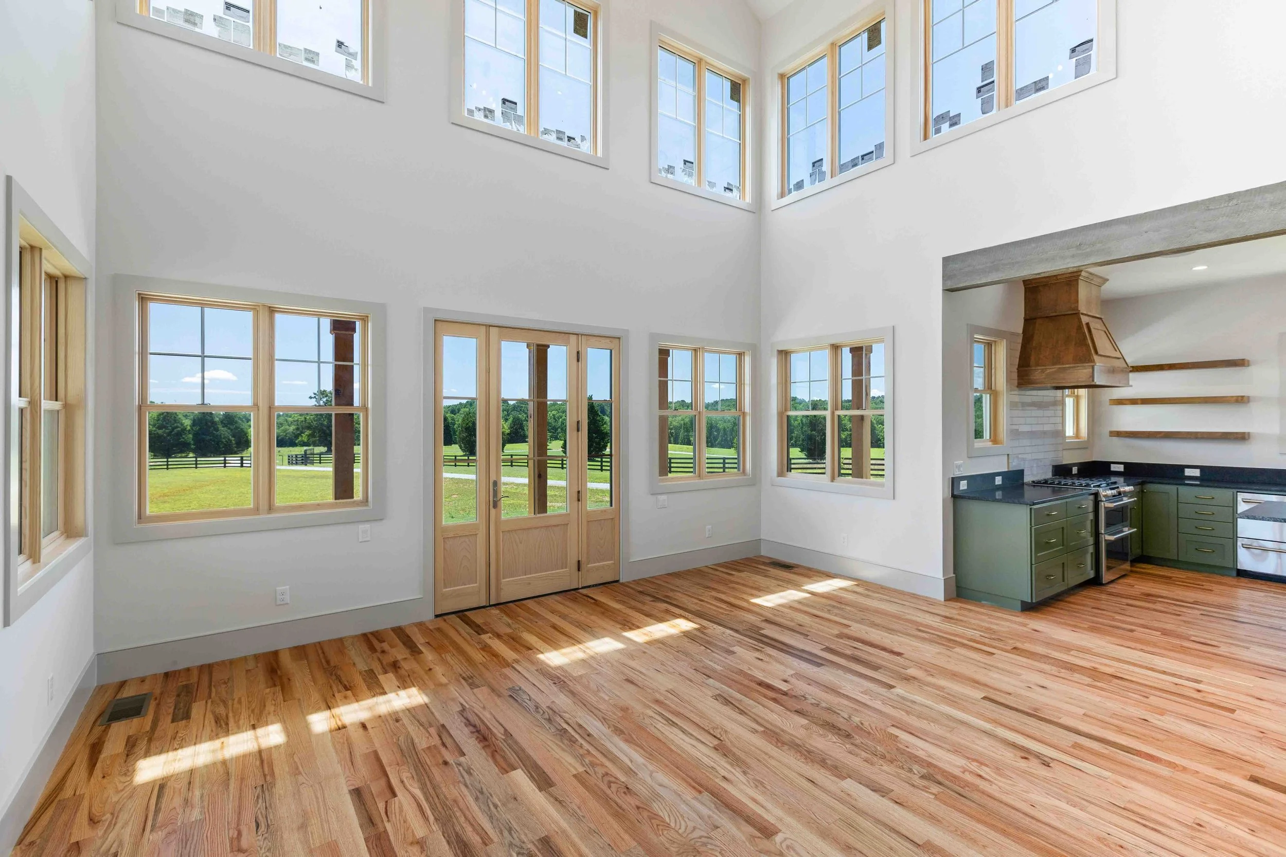 Empty living area with large windows, hardwood floors, and a kitchen corner featuring dark green cabinets and a wooden range hood.