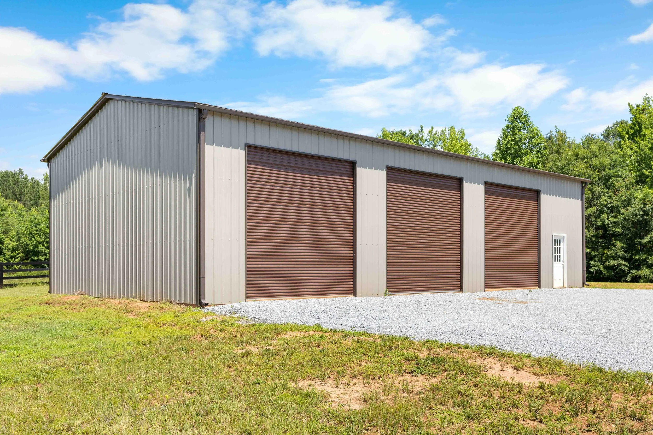 A large metal storage building with three brown roll-up doors and a white door on the side, situated on a gravel and grassy area, with green trees and a blue sky in the background.