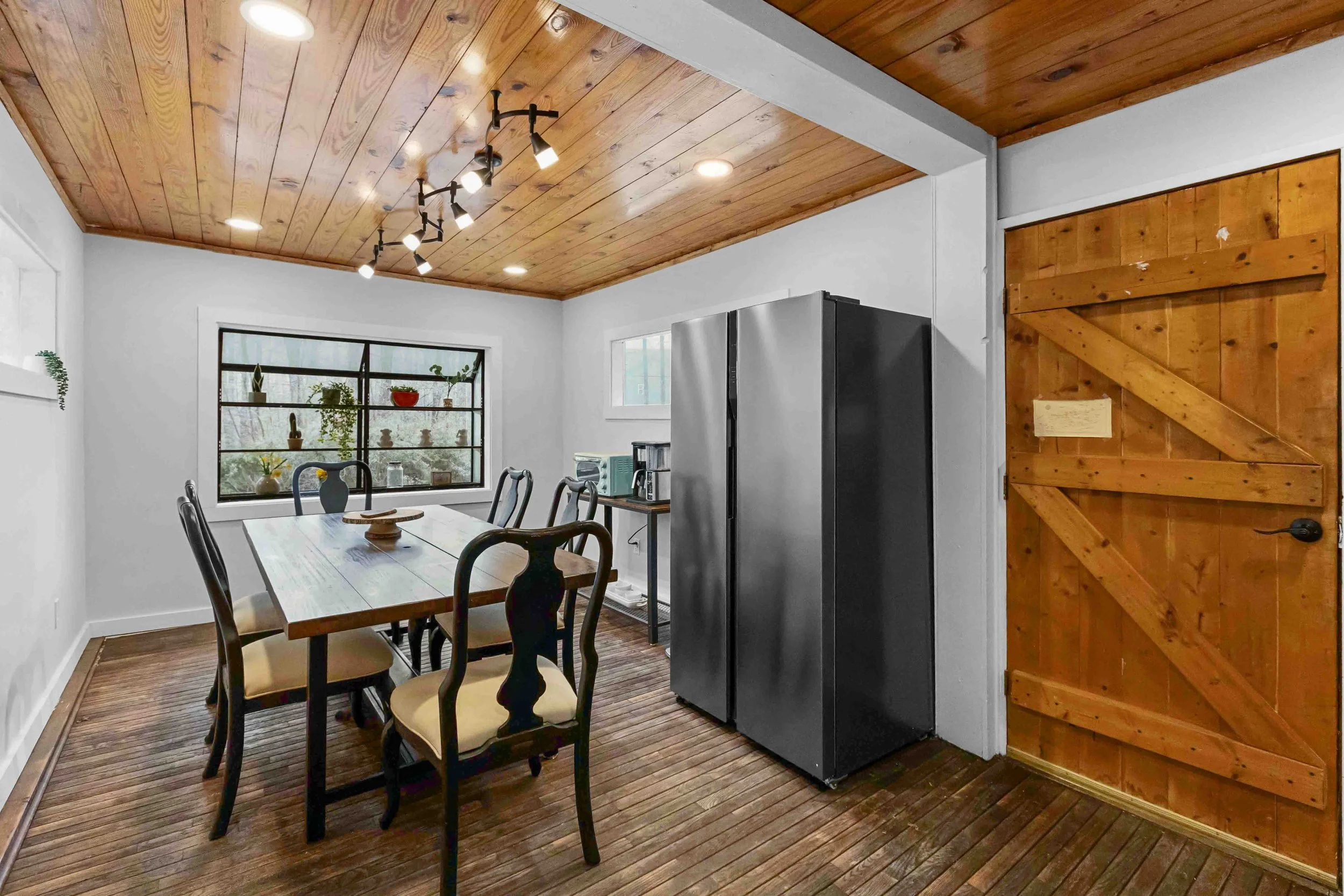 A dining area with a wooden ceiling, white walls, and a wooden floor. There is a rectangular dining table with six chairs around it. A small side table with a microwave and coffee maker is next to a modern stainless steel refrigerator. A large window