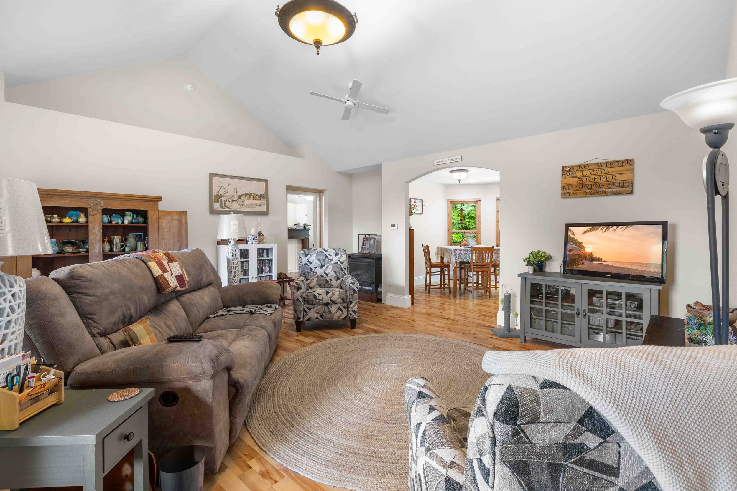 Cozy living room with wooden floors, a round rug, a gray TV stand with a television showing a sunset, patterned armchairs, and a gray sofa with a blanket. Open doorway leading to a dining area with a round table and four chairs, large windows, and a 