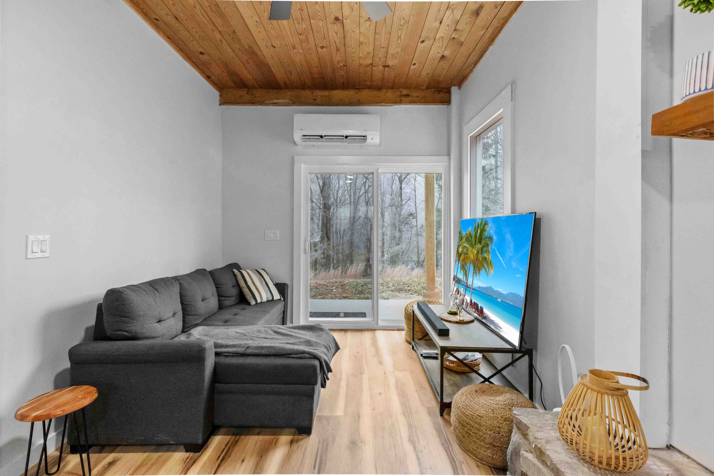 Living room with a gray sectional sofa, window and sliding glass door revealing outdoor trees, a wooden ceiling, a TV on a stand showing a beach scene with palm trees, and various decorative items including a woven basket and wooden lanterns.