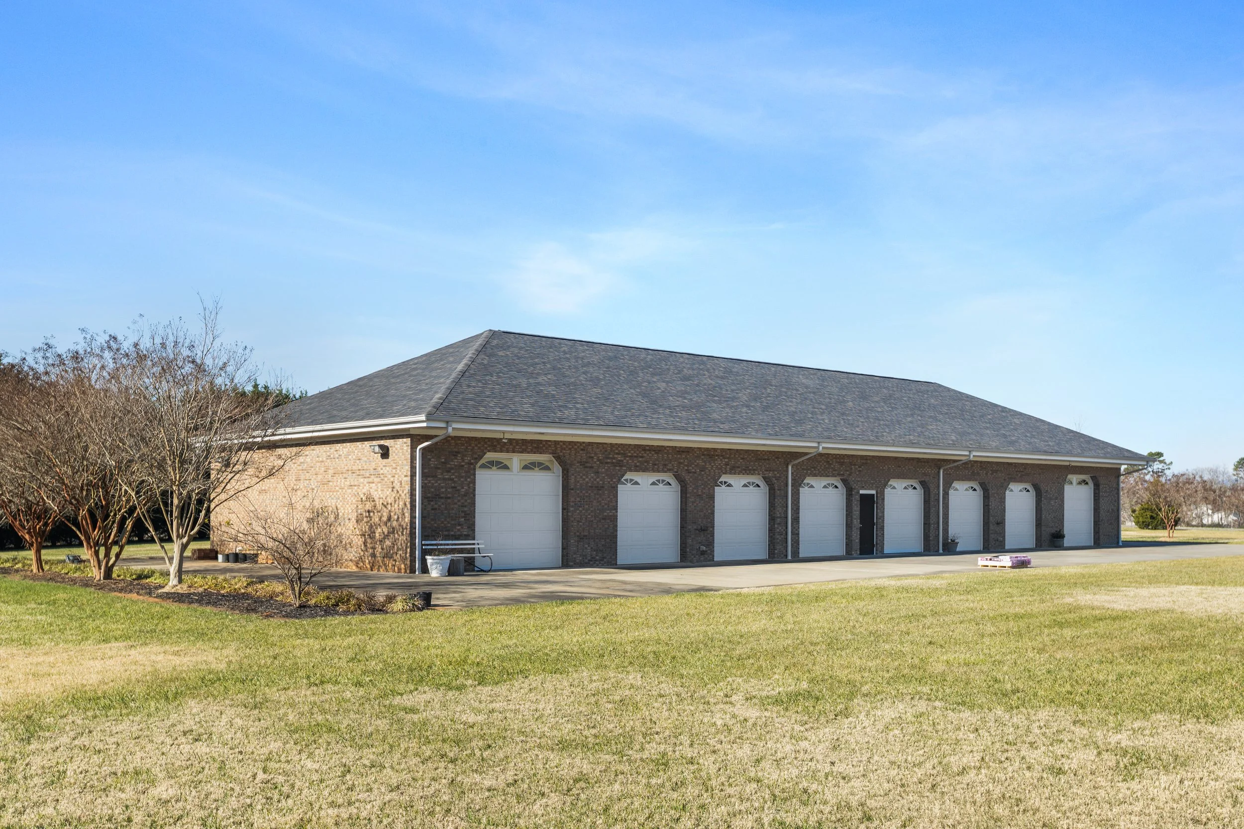 A large brick building with six white garage doors, a black front door, a sloped gray shingle roof, and a lawn with a few trees, under a clear blue sky.
