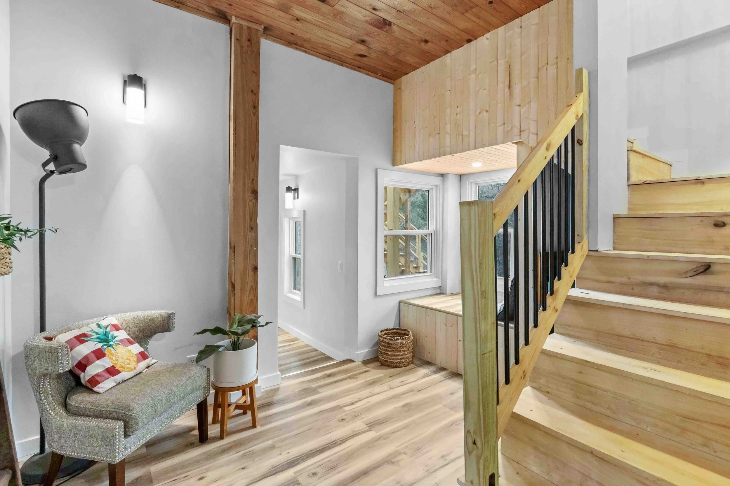 Interior view of a house with wooden staircase, white walls, and natural wood accents, including a small chair with a pineapple pillow, a potted plant, and a window showing outdoor greenery.