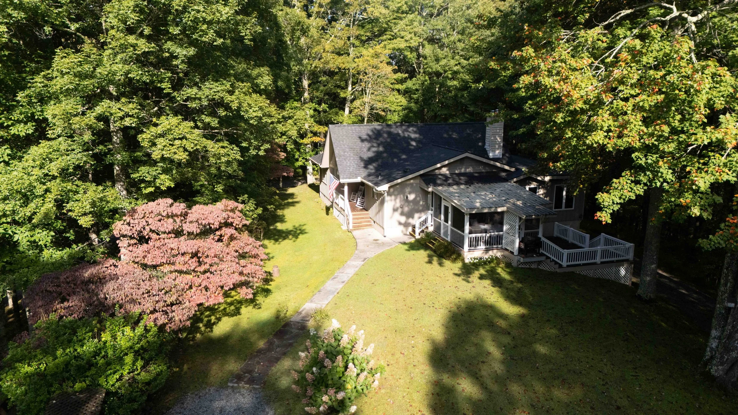 A house surrounded by trees with green and reddish leaves, a grassy yard, and a stone pathway leading to the front steps.