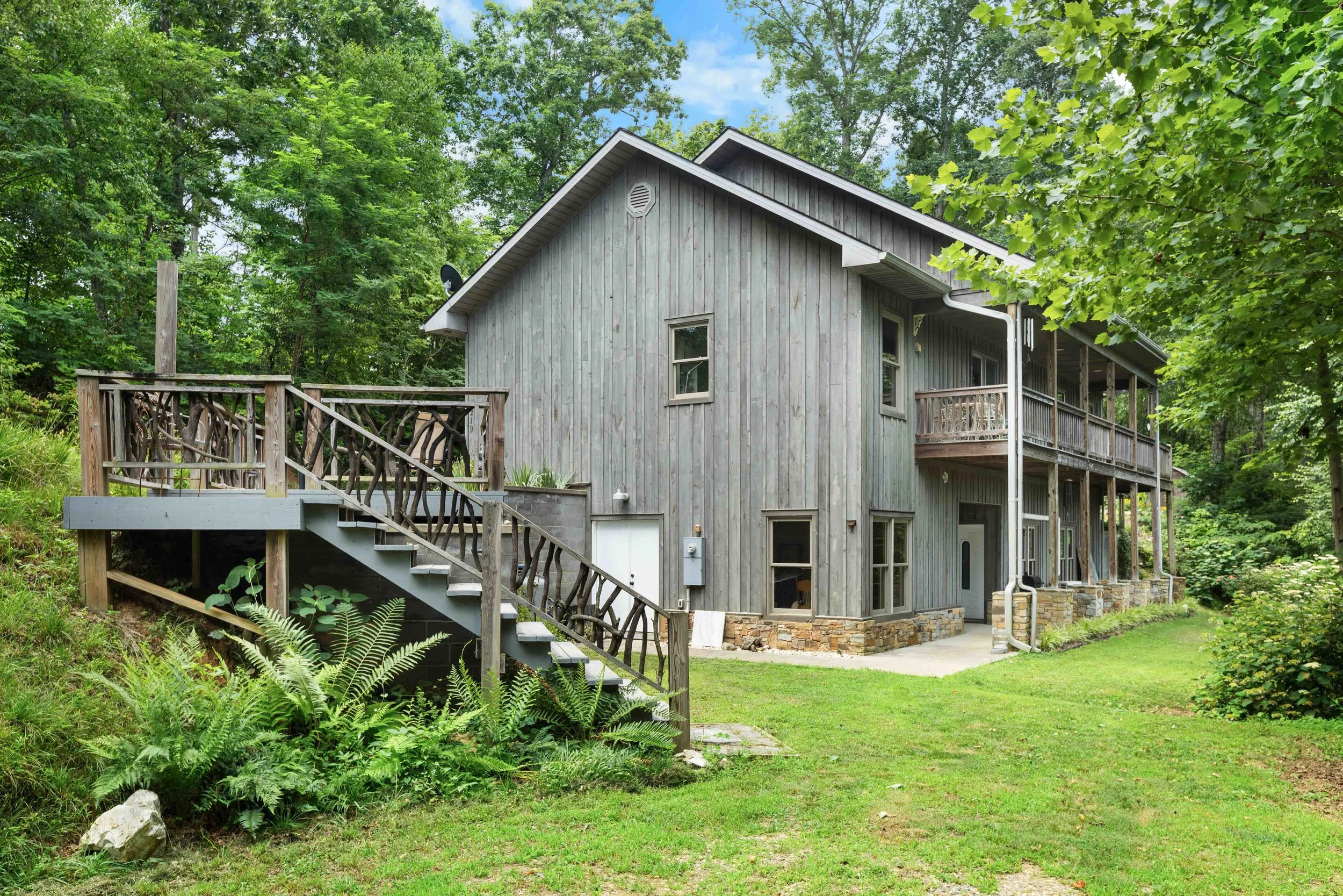A two-story gray wooden house with a balcony on the second floor, surrounded by lush green trees and grass.
