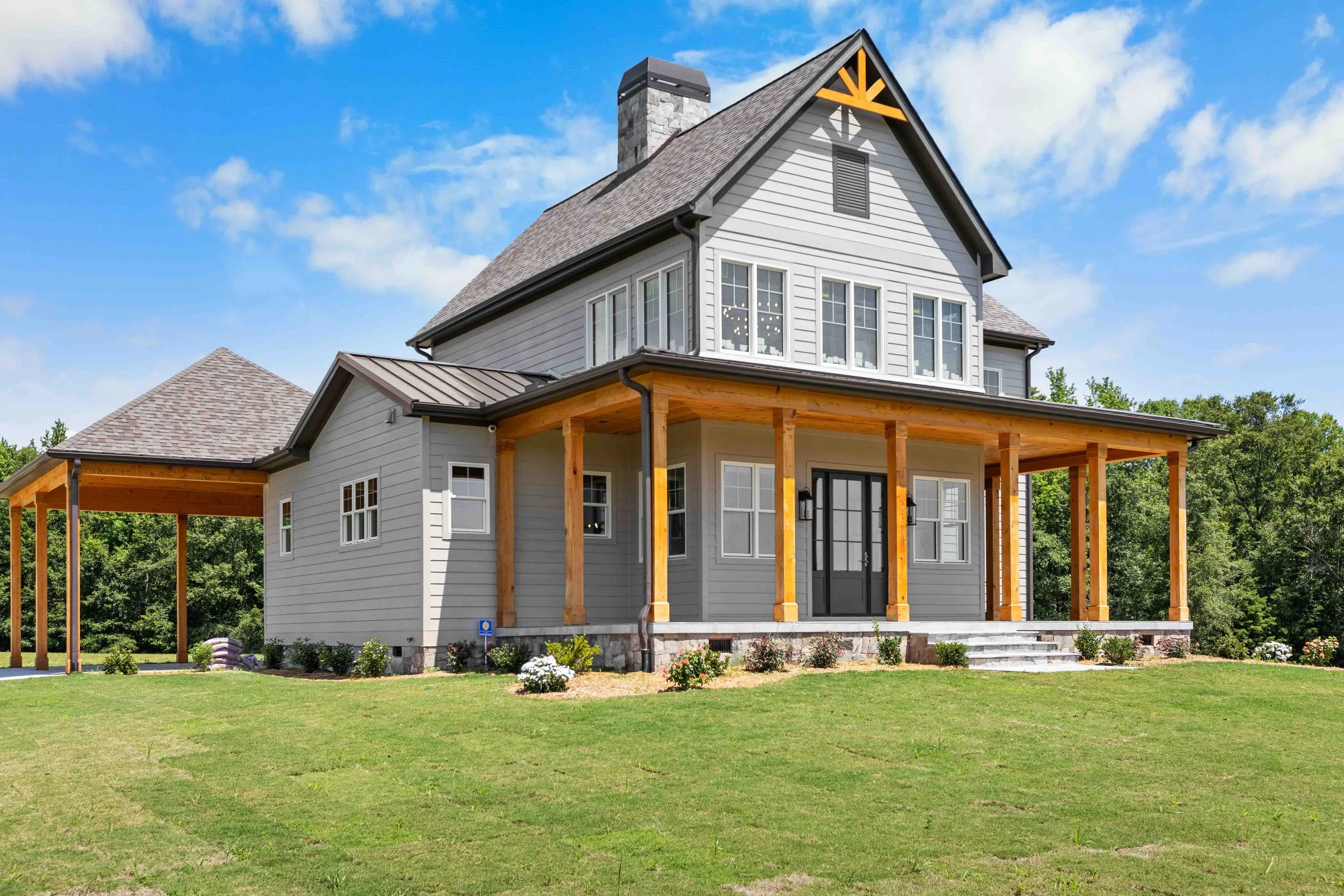 A two-story house with gray siding, black door, wooden porch posts, multiple windows, and a stone chimney, set against a background of trees and a blue sky.