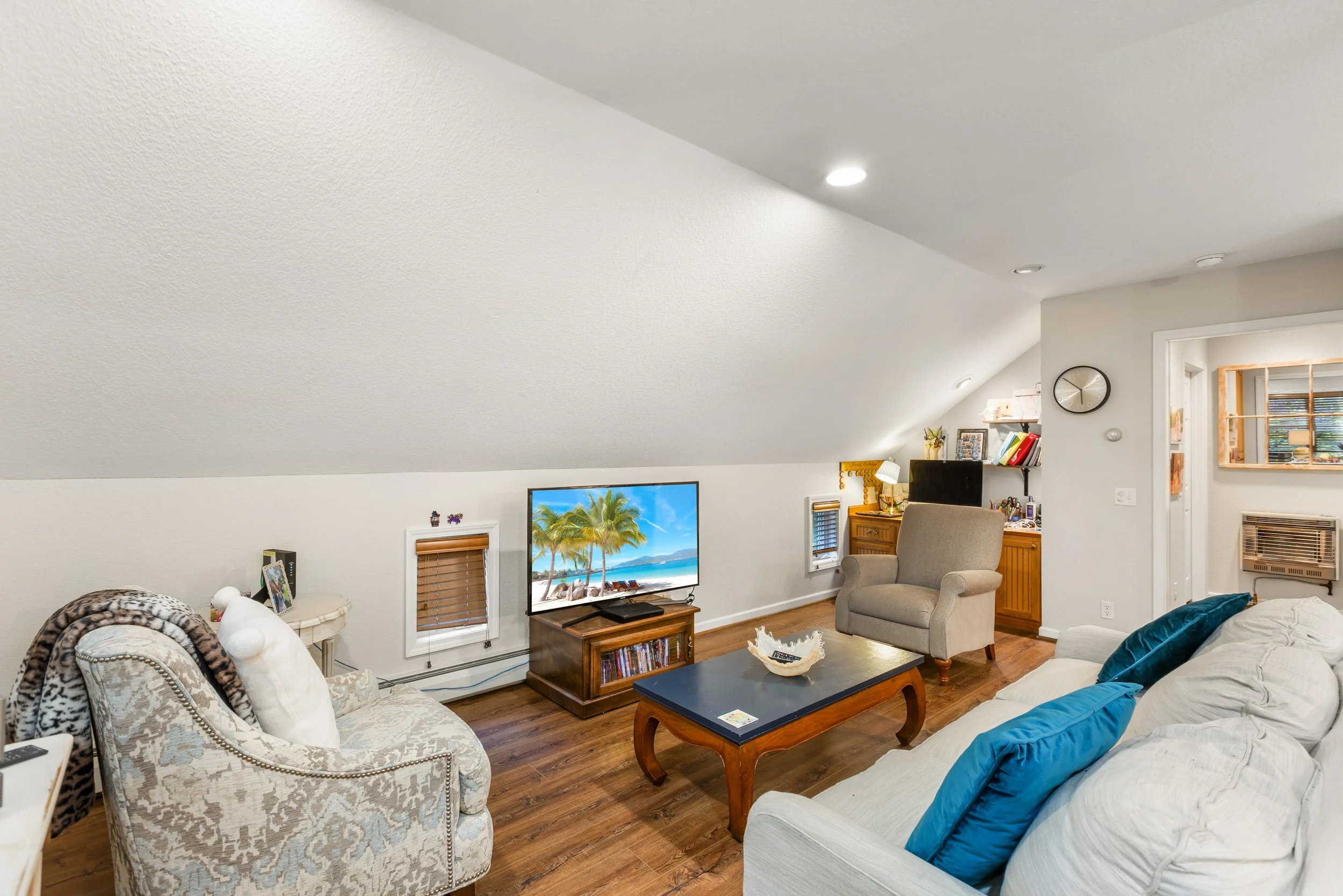 Living room with hardwood floors, beige and white couches, a television, a wooden coffee table, armchair, and various decorations under a white sloped ceiling.