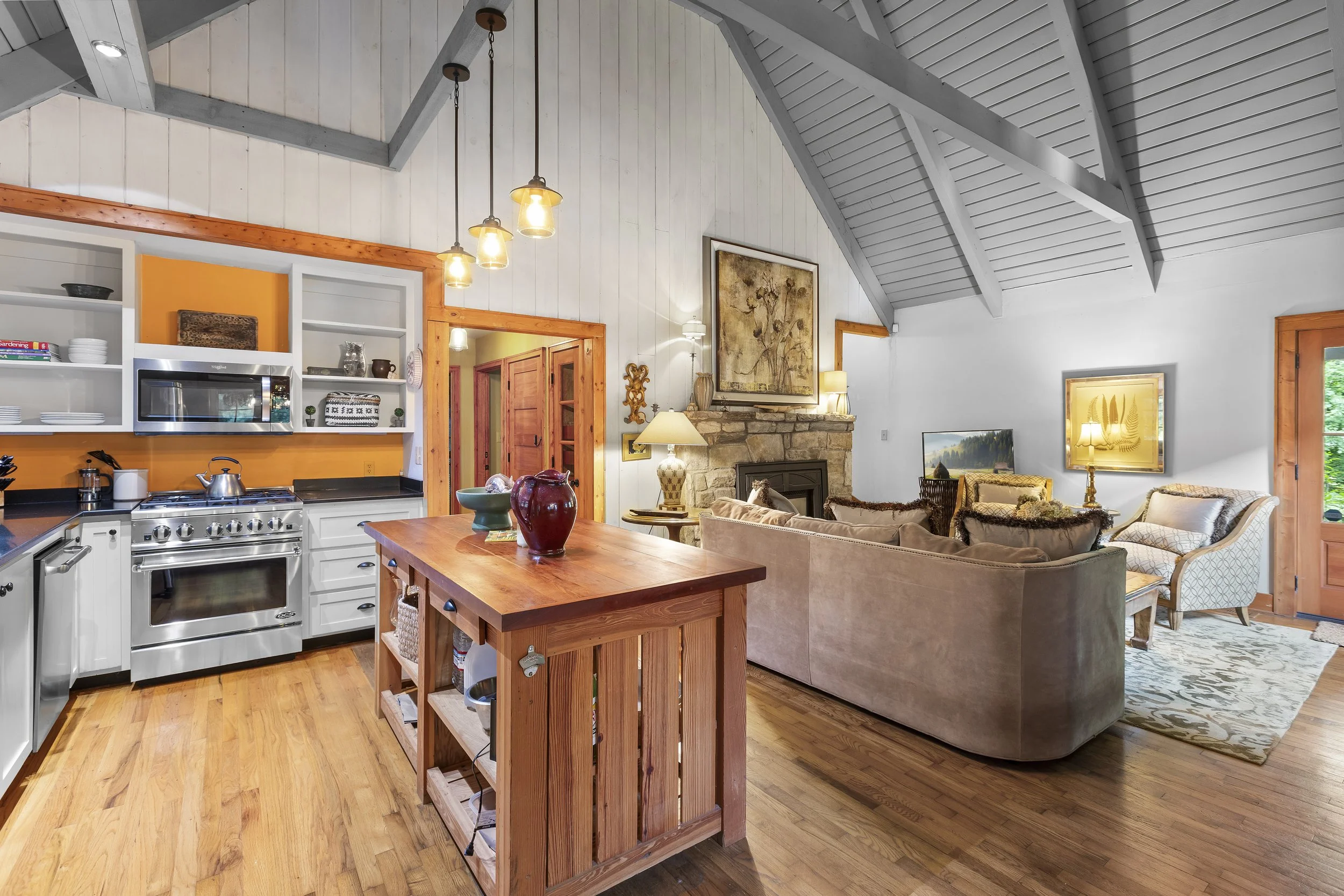 Open-concept living room and kitchen with vaulted ceiling, wood flooring, and rustic decor, including a stone fireplace and a sofa set.