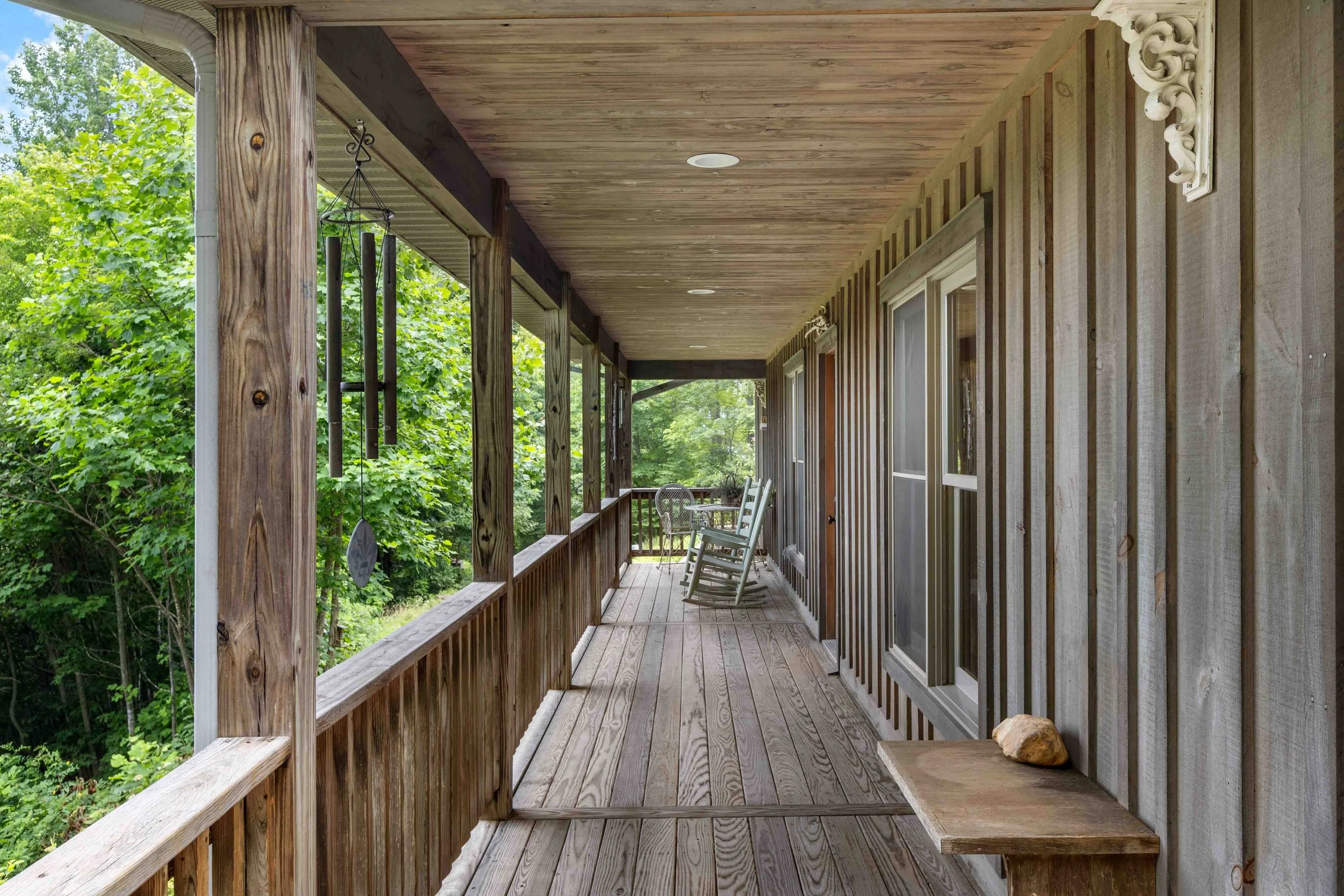 A wooden porch with rocking chairs and a wind chime, surrounded by green trees.