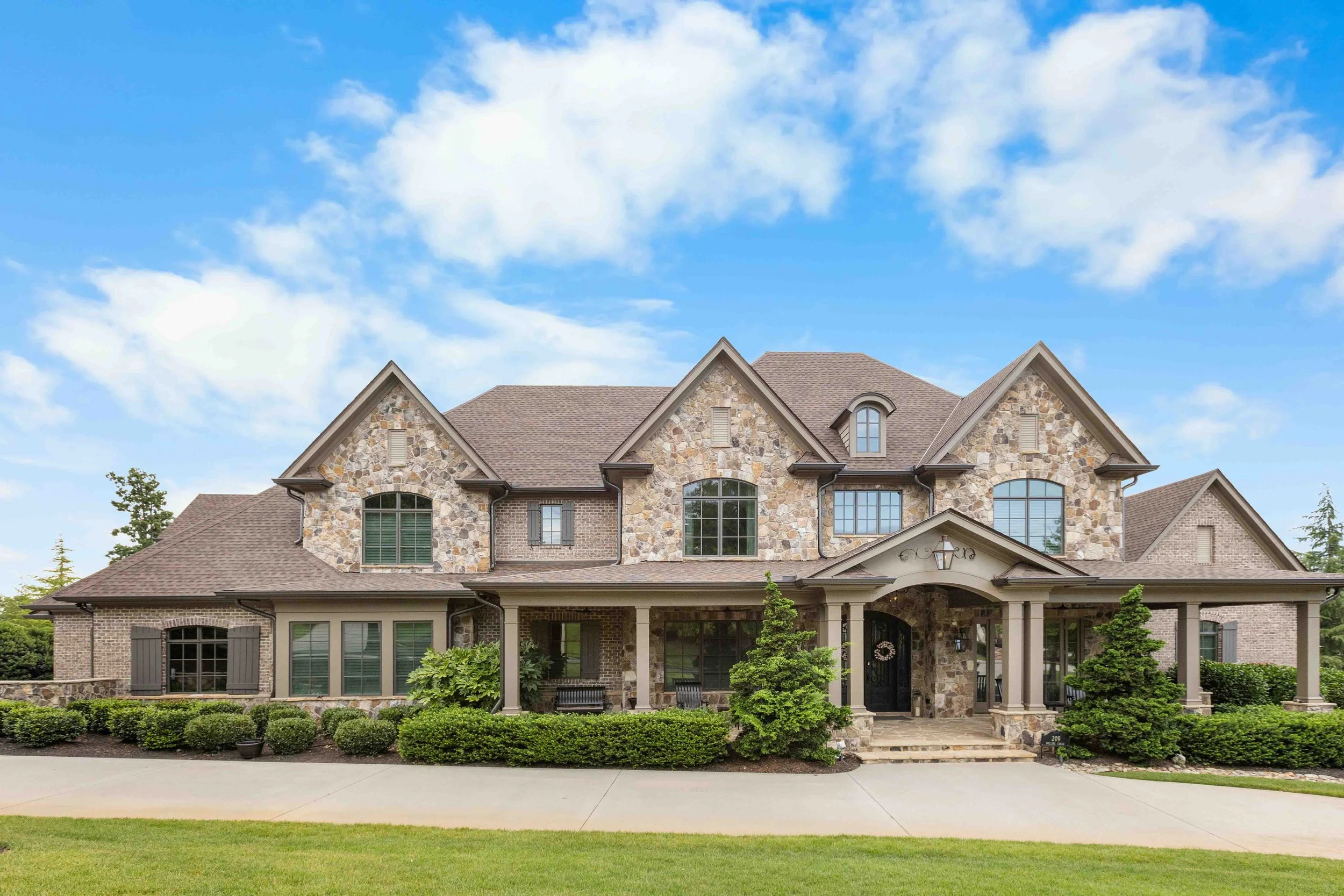 A large two-story house with a stone and brick exterior, multiple gabled roofs, and a front porch with columns, surrounded by neatly trimmed bushes and a green lawn under a partly cloudy blue sky.
