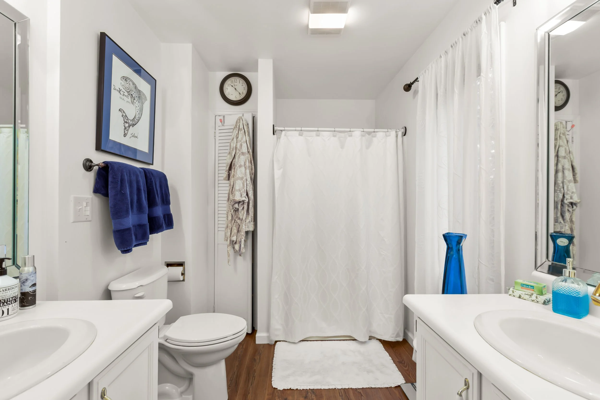 A bathroom with white walls, a white shower curtain, a bathroom sink, a toilet, a mirror, two blue towels, a clock, a framed fish print, and a white rug on a wooden floor.