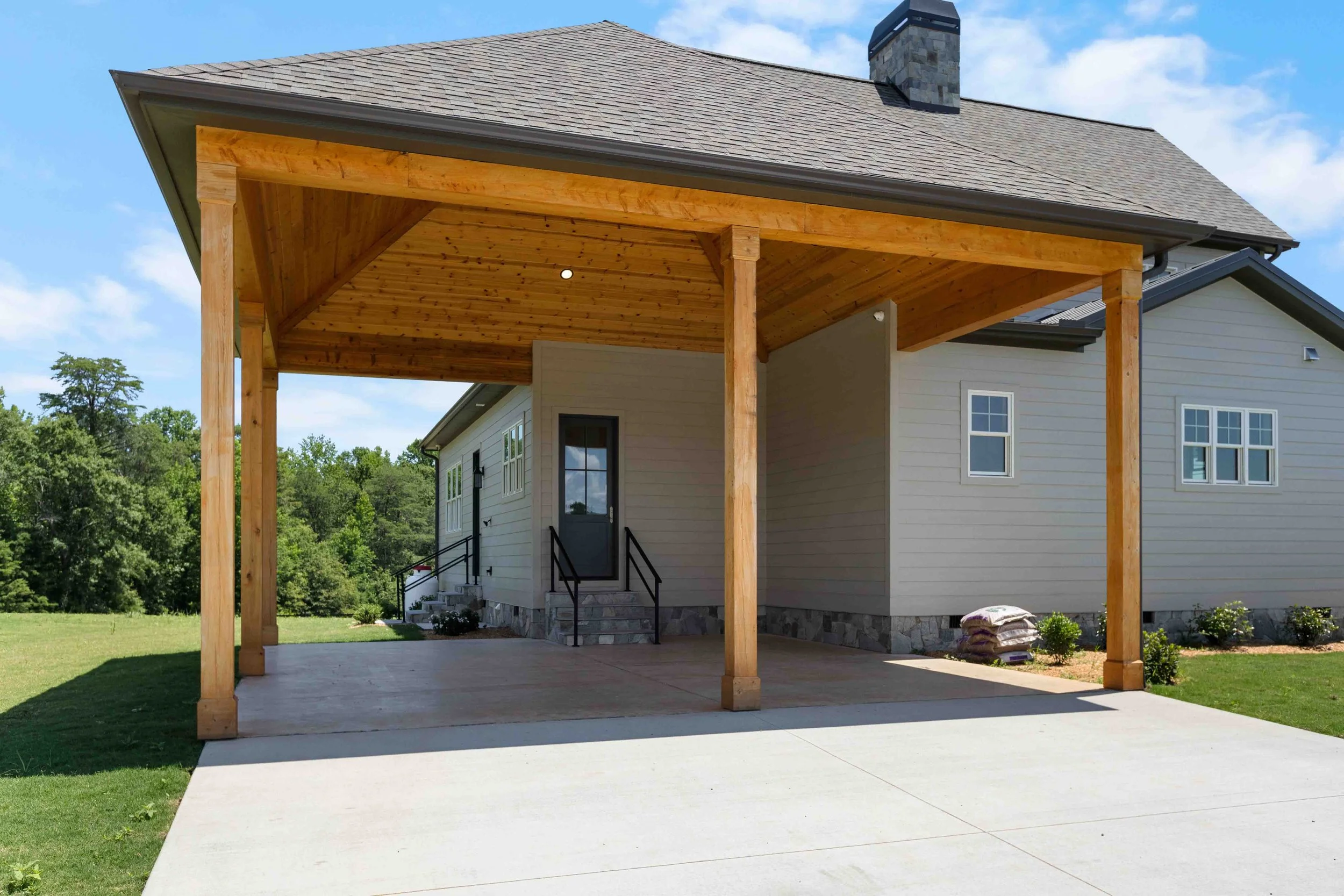 Back view of a house with a wooden porch structure supported by four large wooden beams, with a concrete driveway leading up to it, surrounded by grass and trees.