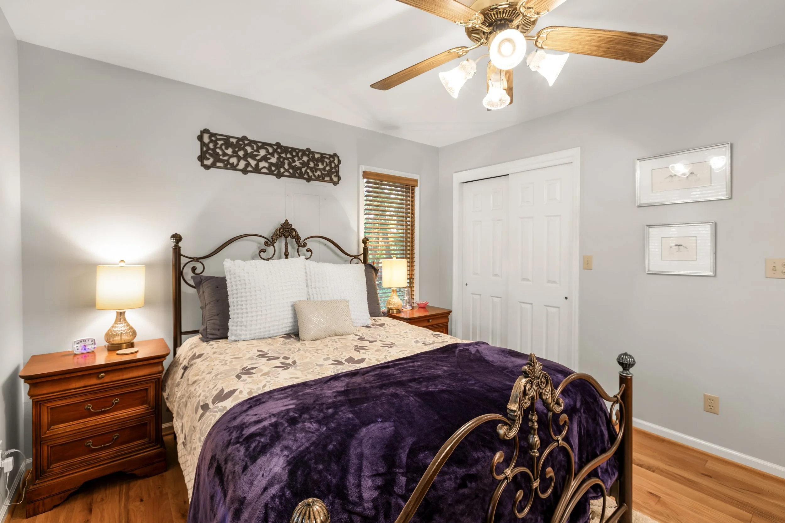 A bedroom with a brass bed, dressed in a floral quilt and purple throw blanket. Two nightstands with lamps are on either side of the bed. There are window blinds, and framed pictures on the wall.
