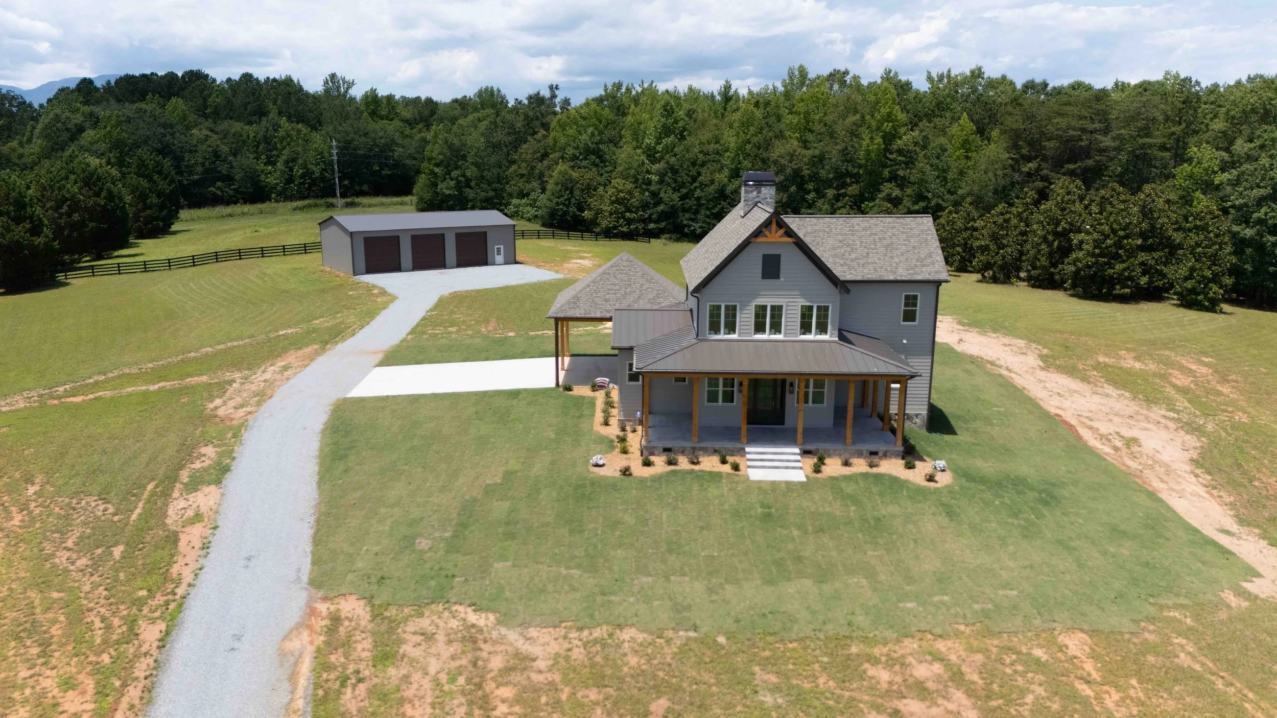 A house with a gray exterior and front porch, situated on a spacious grassy yard with a gravel driveway leading to a detached garage, surrounded by green trees and hills.