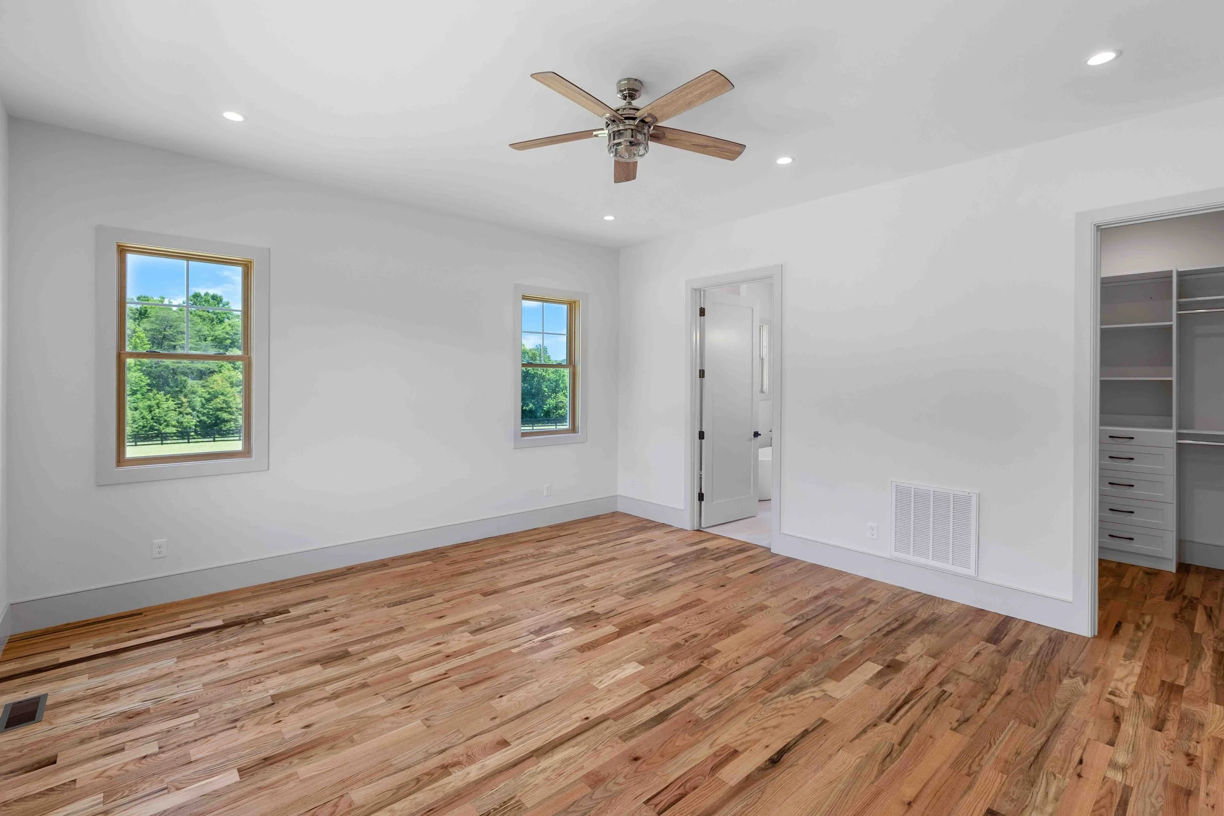 Empty room with hardwood floors, white walls, two windows, a ceiling fan, and an open closet.