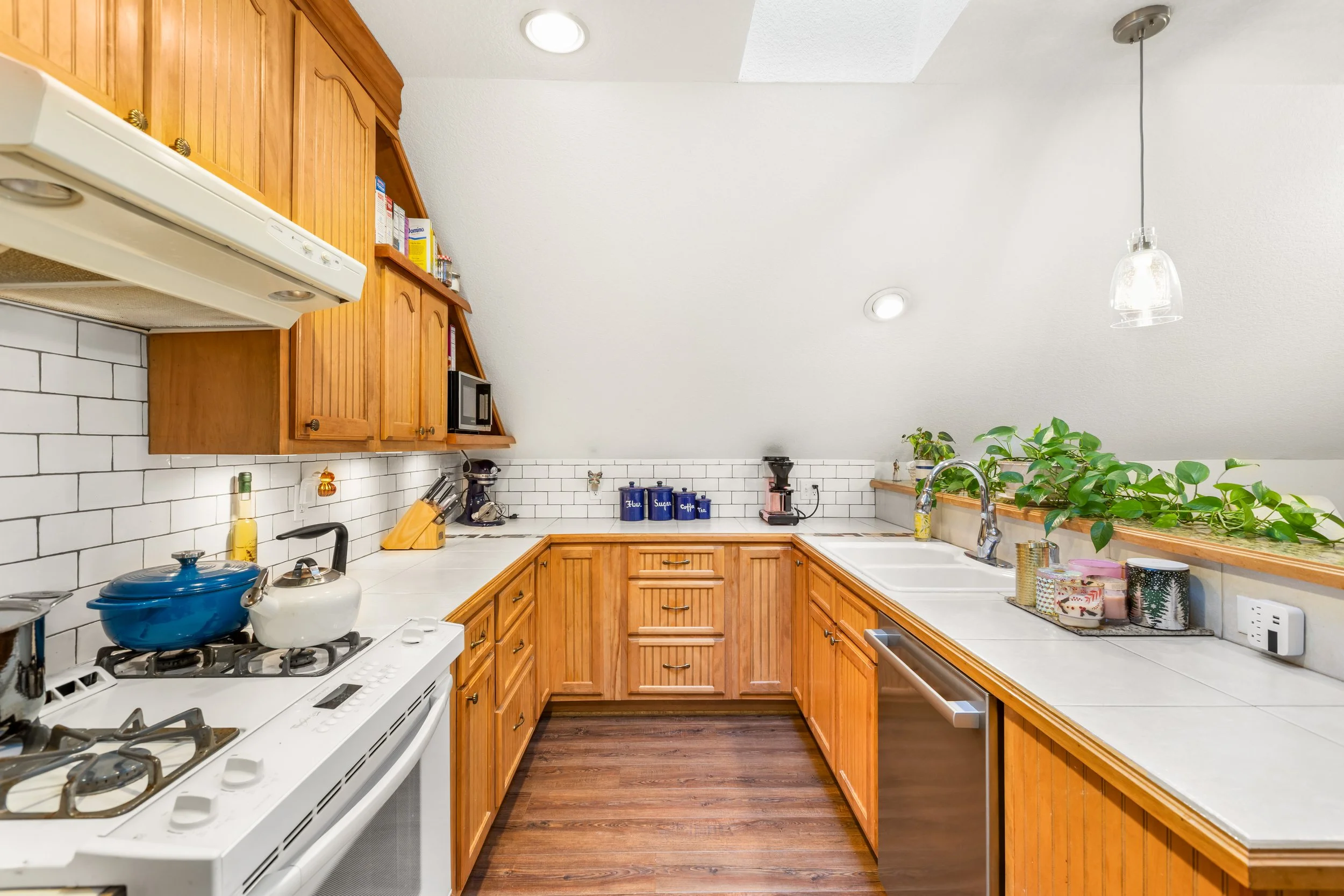 Kitchen with white countertops, wooden cabinets, white subway tile backsplash, and kitchen appliances including a stove with pots, toaster, microwave, and coffee maker. There are green plants along the window ledge and a hanging glass light.