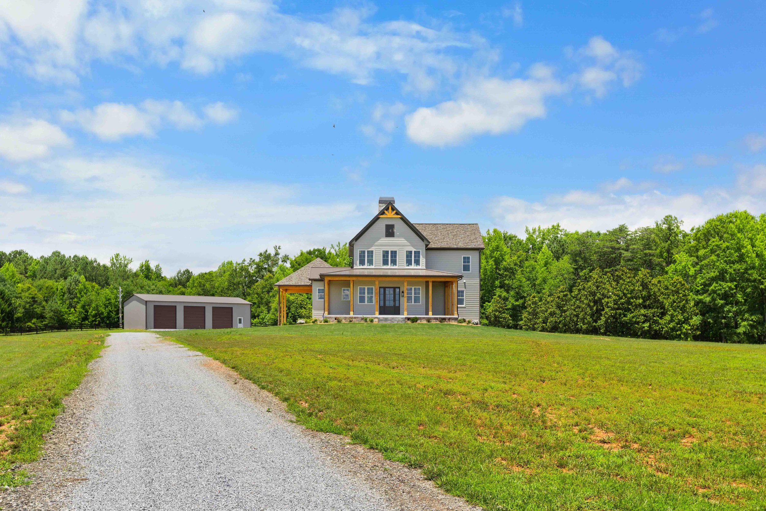 A two-story house with a front porch and attached garage, set on a grassy hill with a gravel driveway, surrounded by green trees under a partly cloudy blue sky.