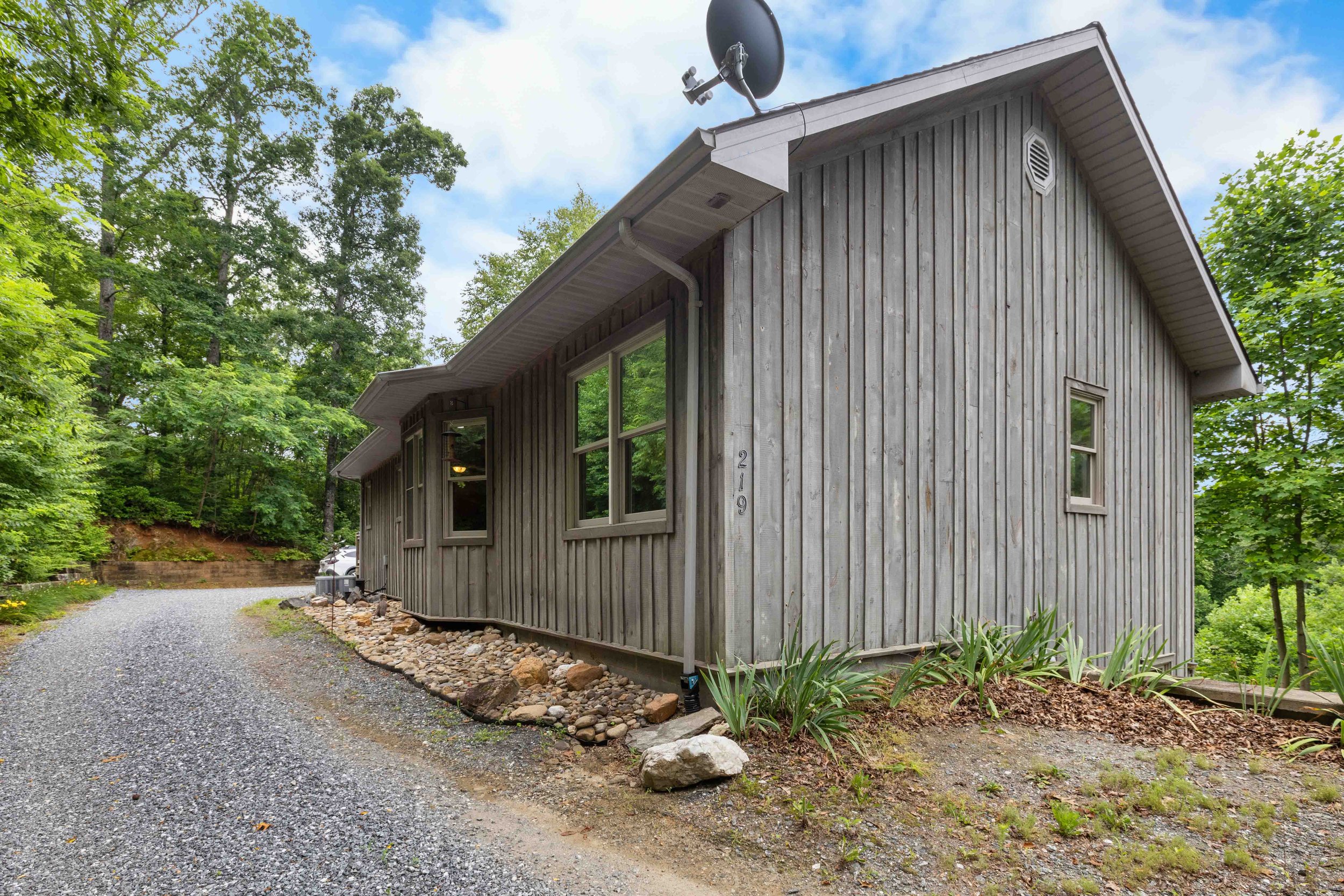 Gray wooden house with a gravel driveway, several windows, a satellite dish on the roof, surrounded by green trees and plants.