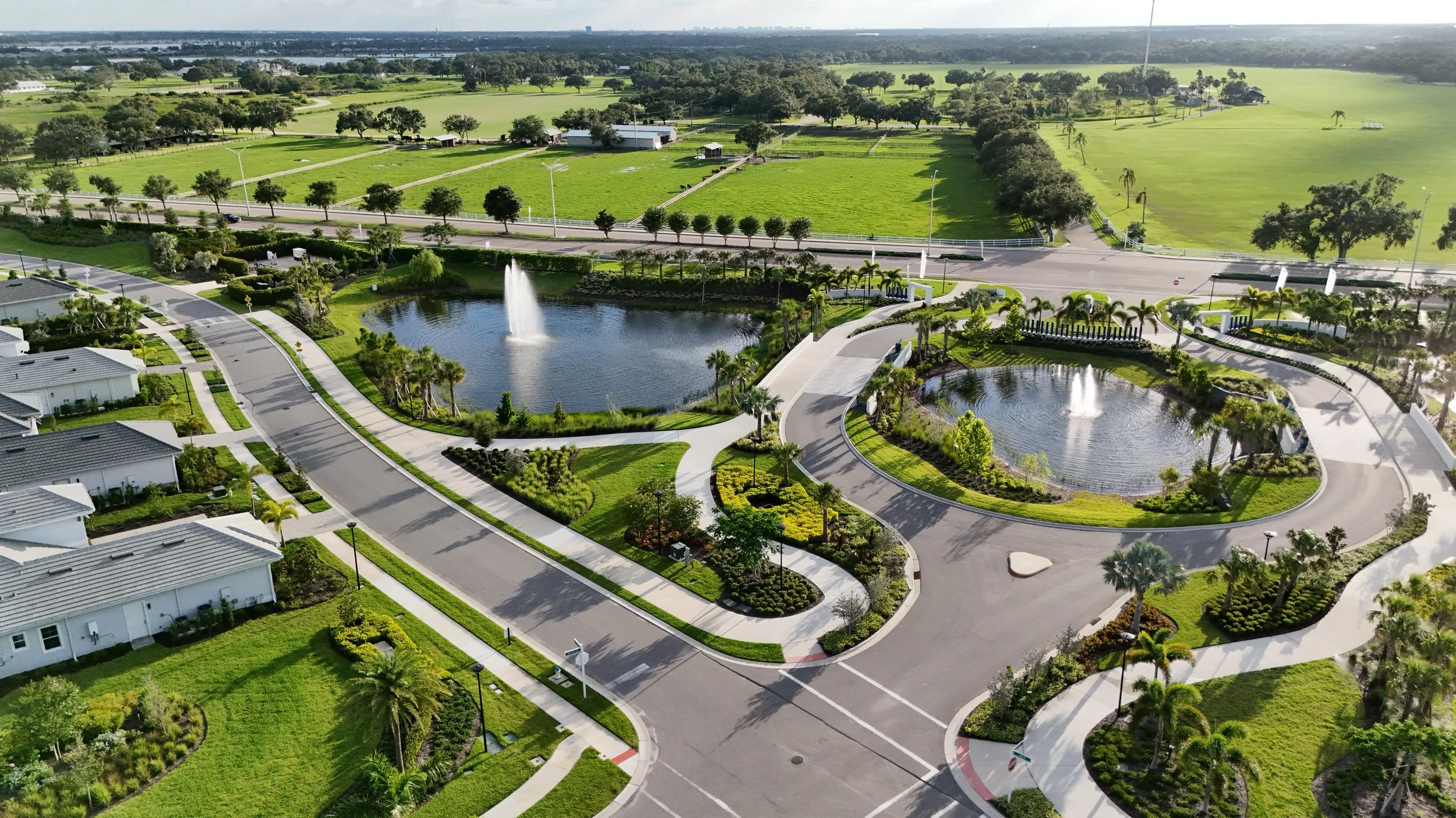 Aerial view of a residential neighborhood with green lawns, palm trees, and two circular ponds with fountains, surrounded by sidewalks and roads, with open green fields and trees in the background.