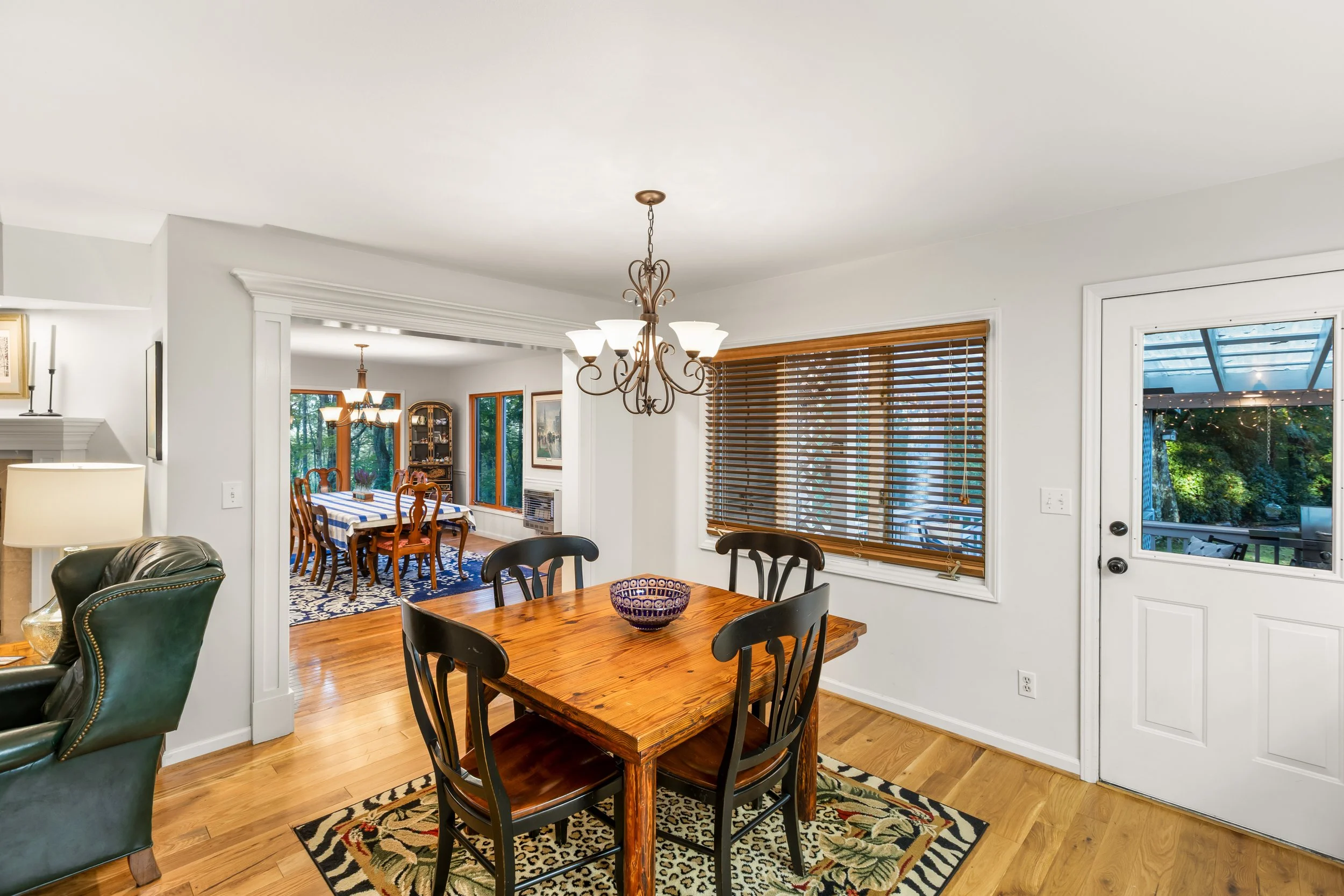 Dining area with wooden table and four black chairs, window with blinds, and a door leading outside to a patio with greenery.