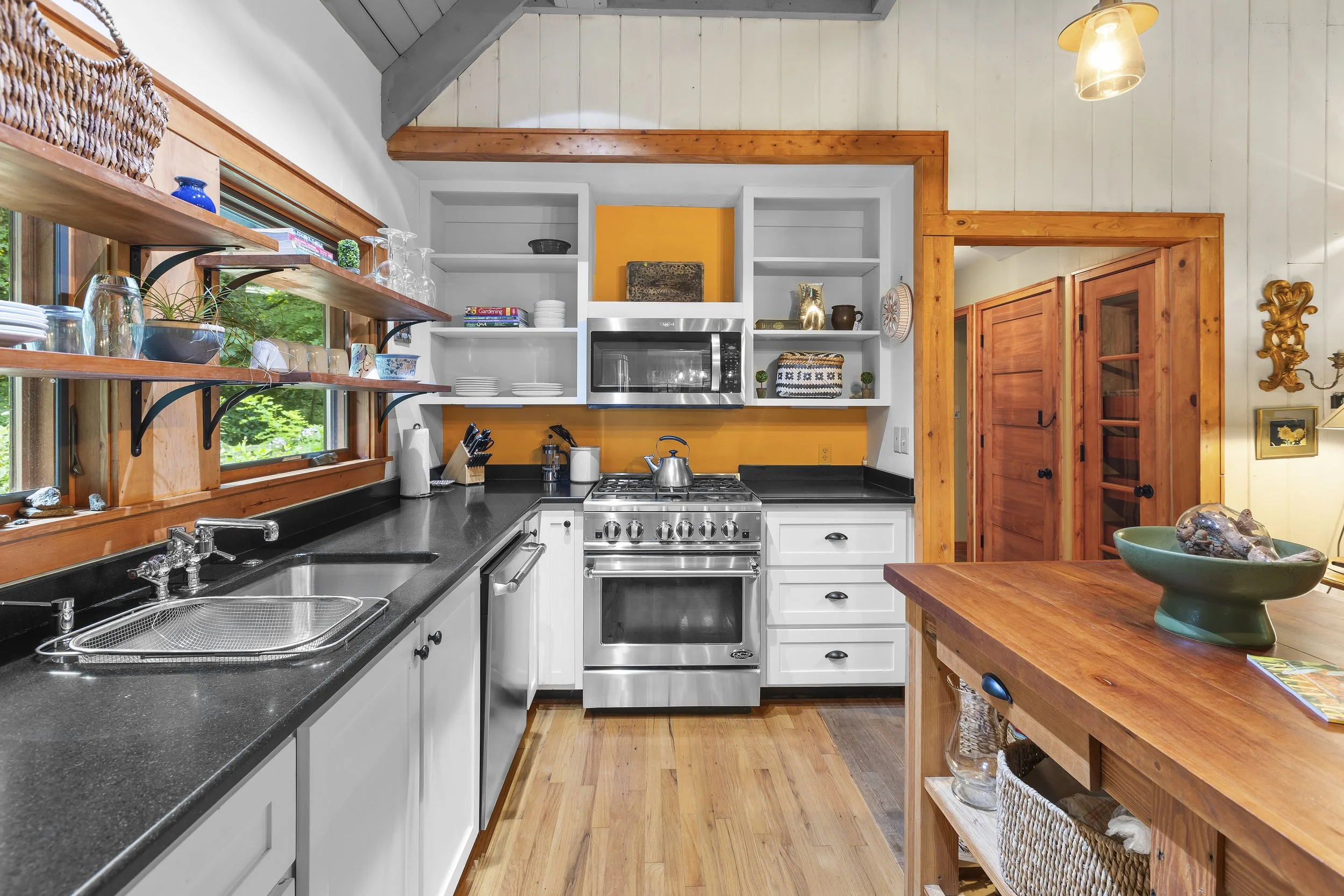 Kitchen with white cabinets, black countertops, and wooden accents, including a wooden island, open shelving with dishes, and a bright yellow backsplash behind a stainless steel stove and microwave. Windows with wood frames reveal green outside folia