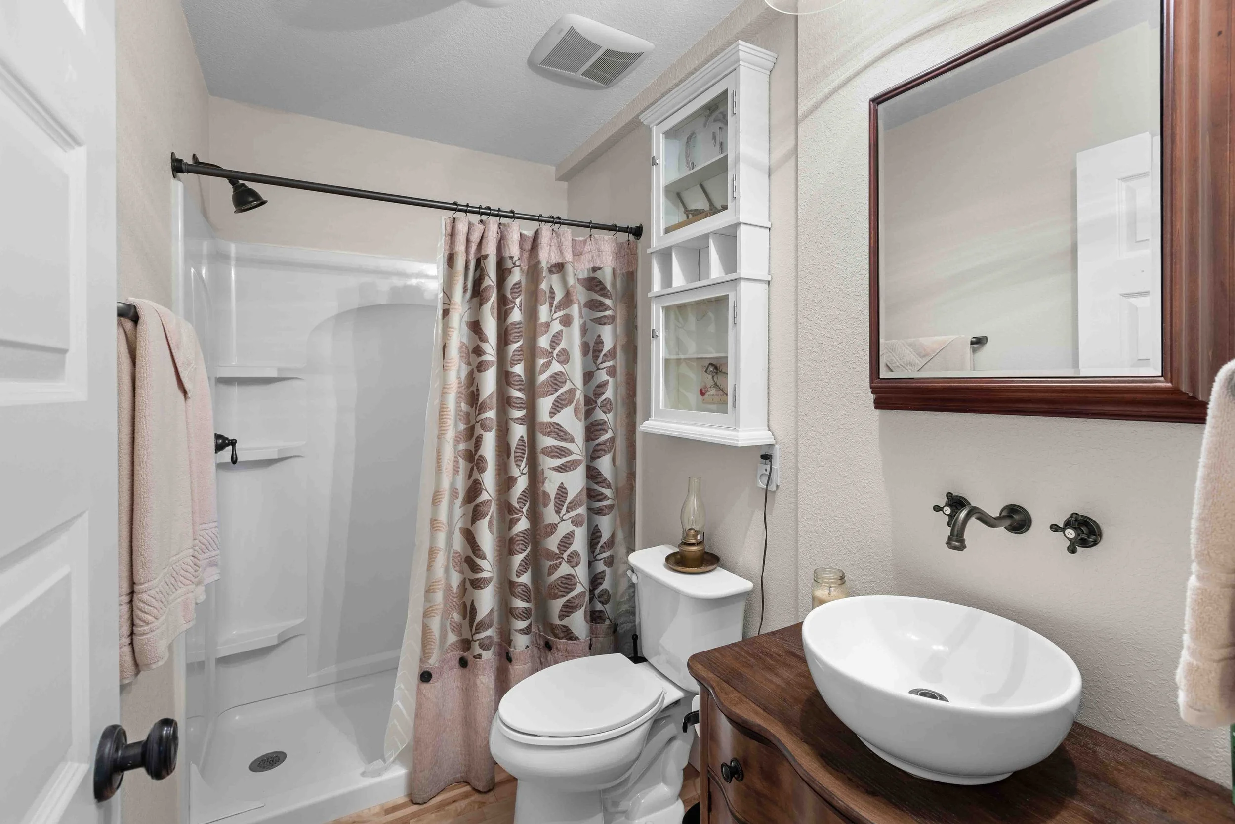Small bathroom with a shower stall with a curtain, a white toilet, a wooden vanity with a round vessel sink, a rectangular mirror with a dark wooden frame, and wall-mounted faucet and handles.