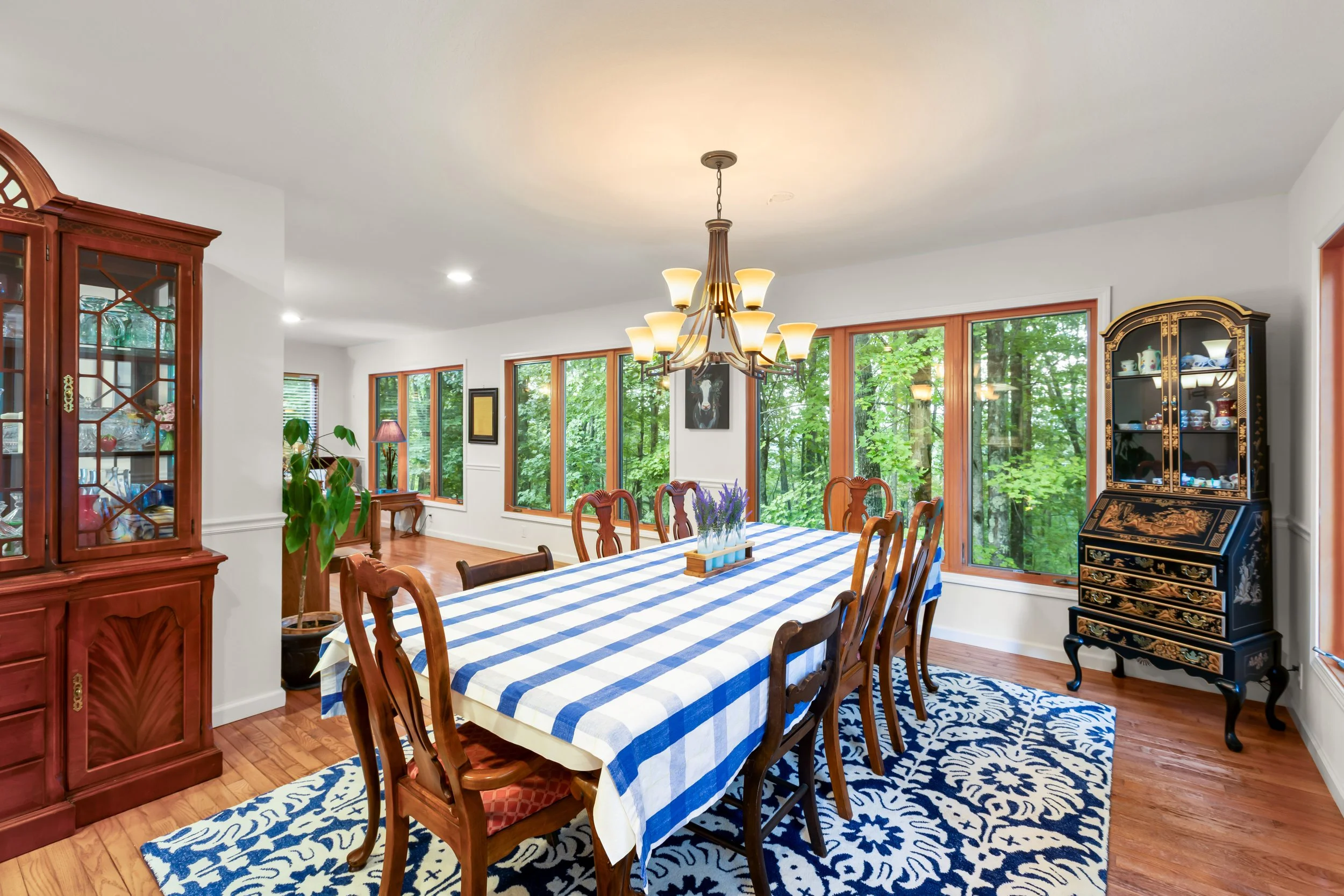 Bright dining room with a long table covered in a blue and white checkered tablecloth, surrounded by wooden chairs, accessing large windows with a view of green trees outside. Features a black and gold china cabinet, a wooden hutch, a blue and white 