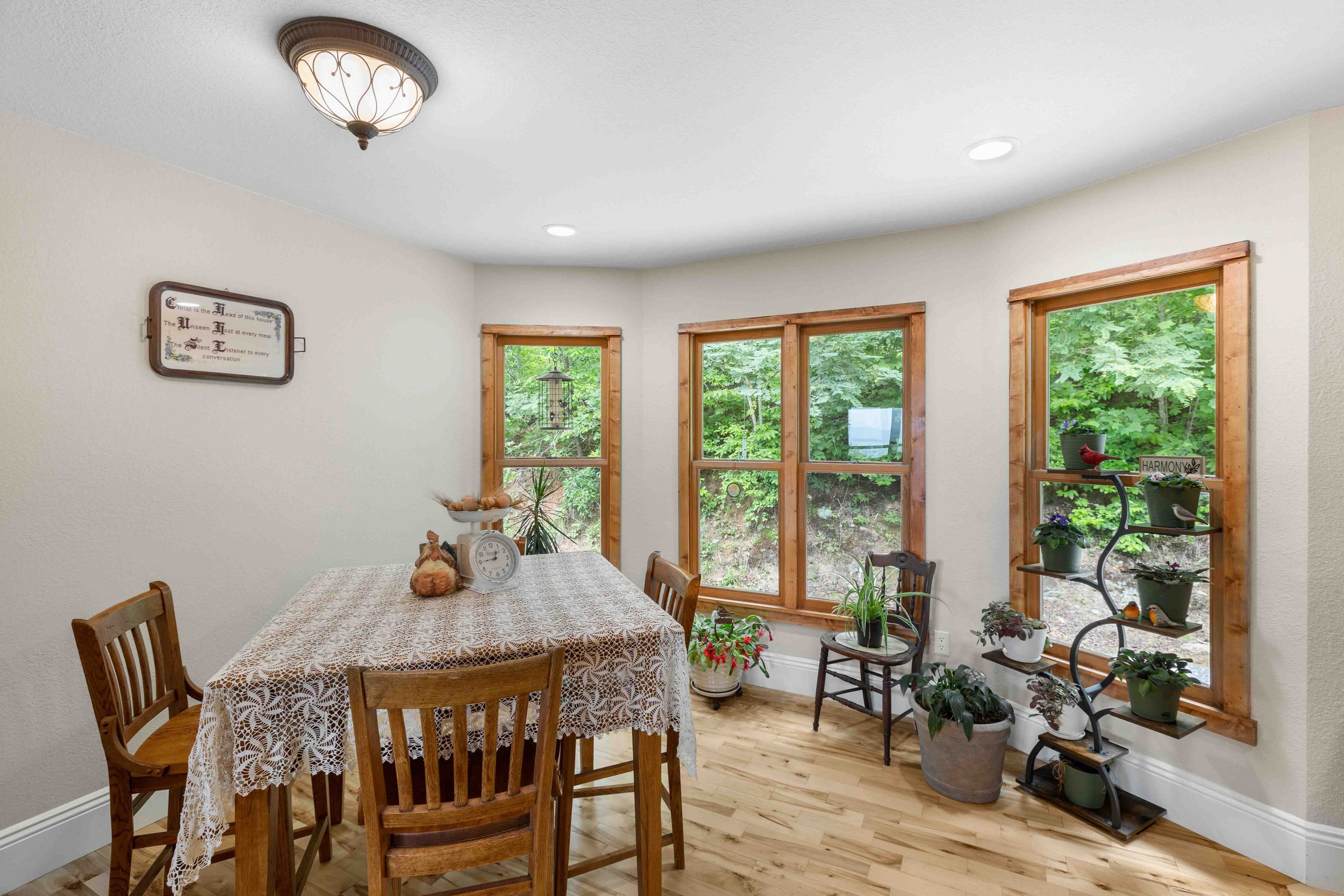 A bright dining room with a wooden table covered by a lace tablecloth, surrounded by four wooden chairs. There are three large windows with wood trim, showing green trees outside. Potted plants are on a black metal shelf by the window and on the floo