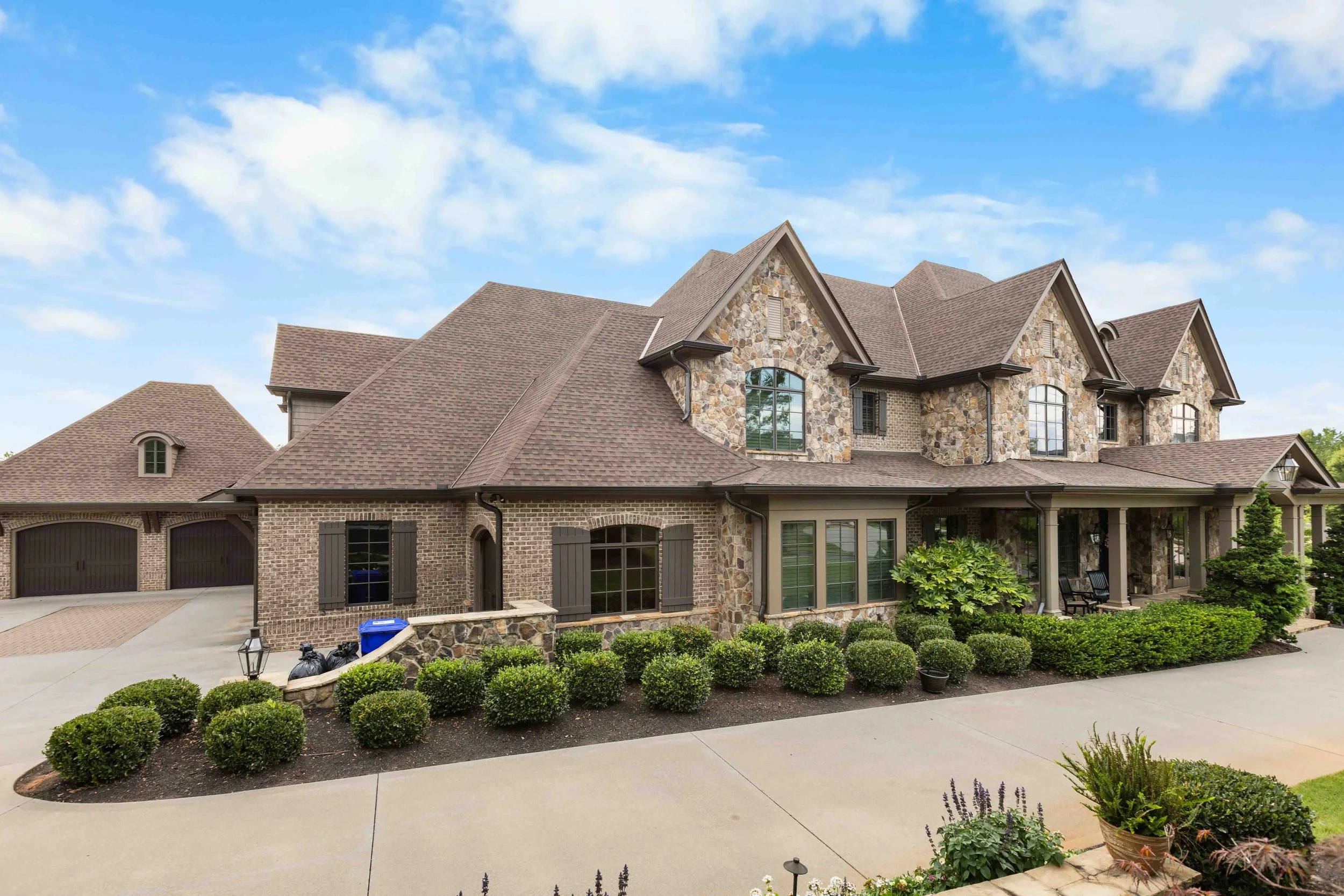 Large, upscale house with stone and brick exterior, multiple gable roofs, a front porch with chairs, landscaped bushes, and a driveway leading to a three-car garage under a partly cloudy sky.